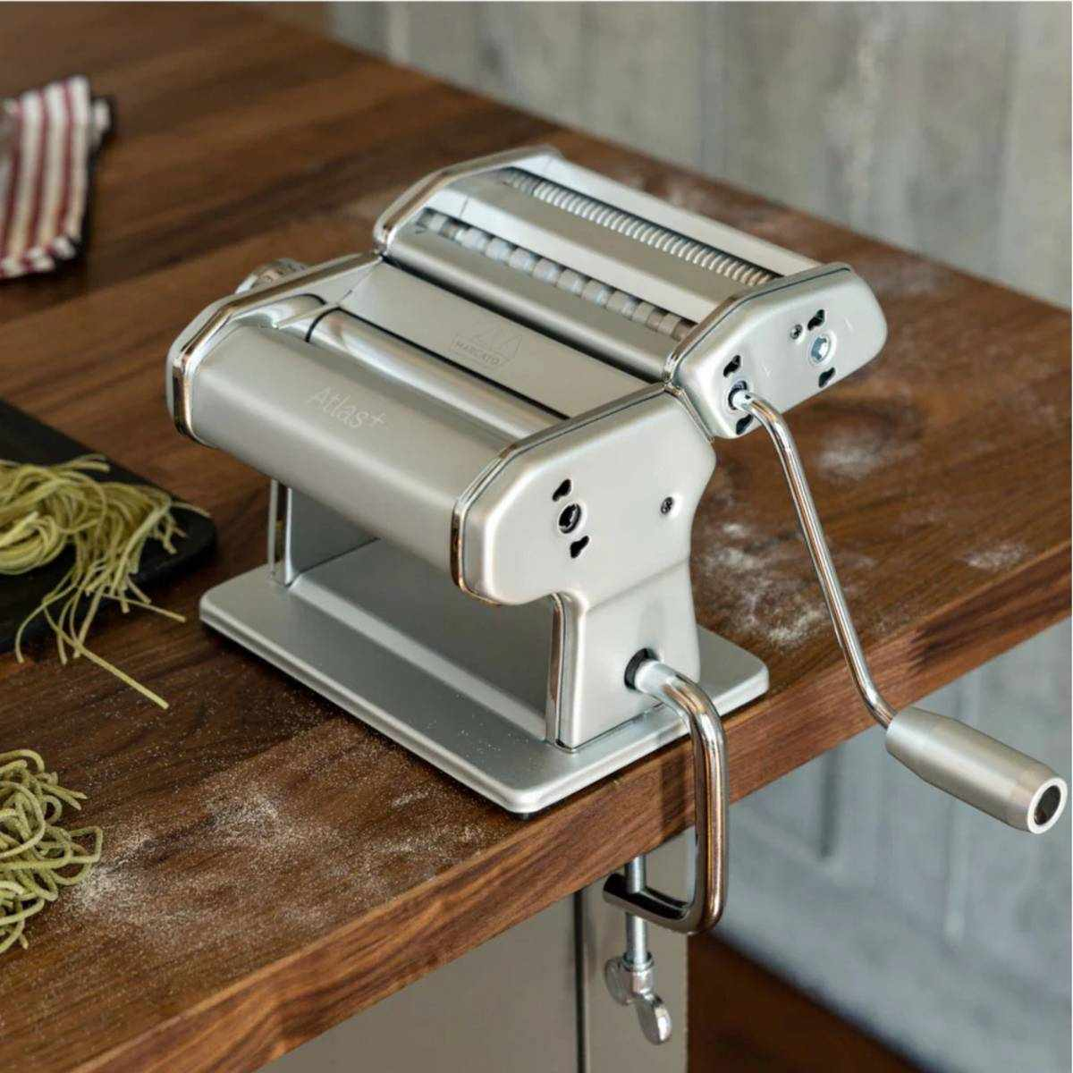 Eine silberne Nudelmaschine mit Handkurbel steht auf einem Holztisch.
