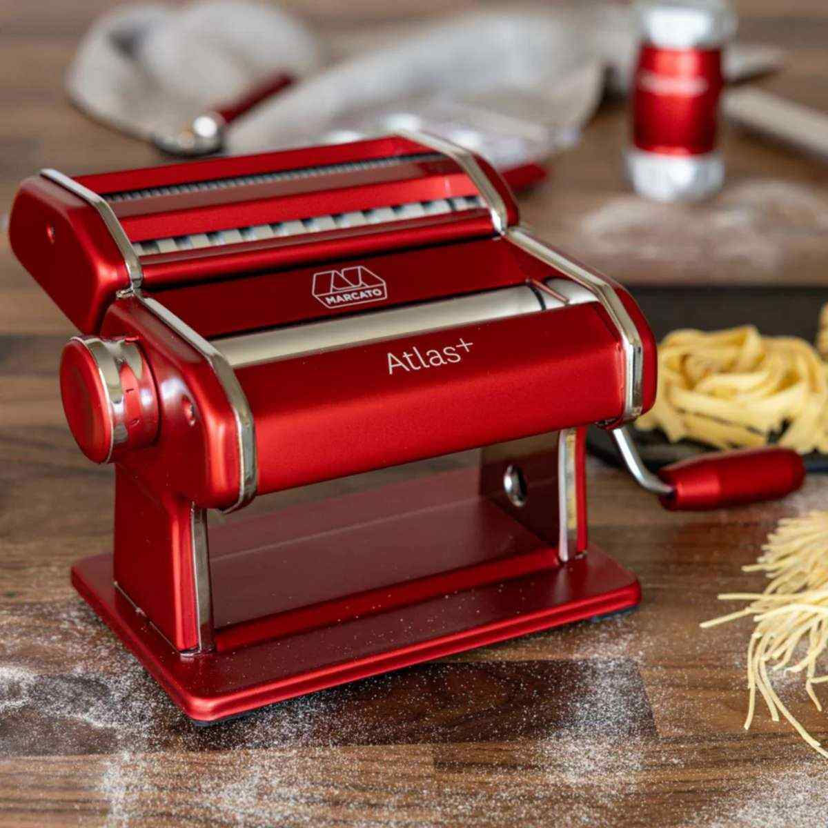 Rote Nudelmaschine mit Silberakzenten auf einem Holztisch, daneben Nudeln.