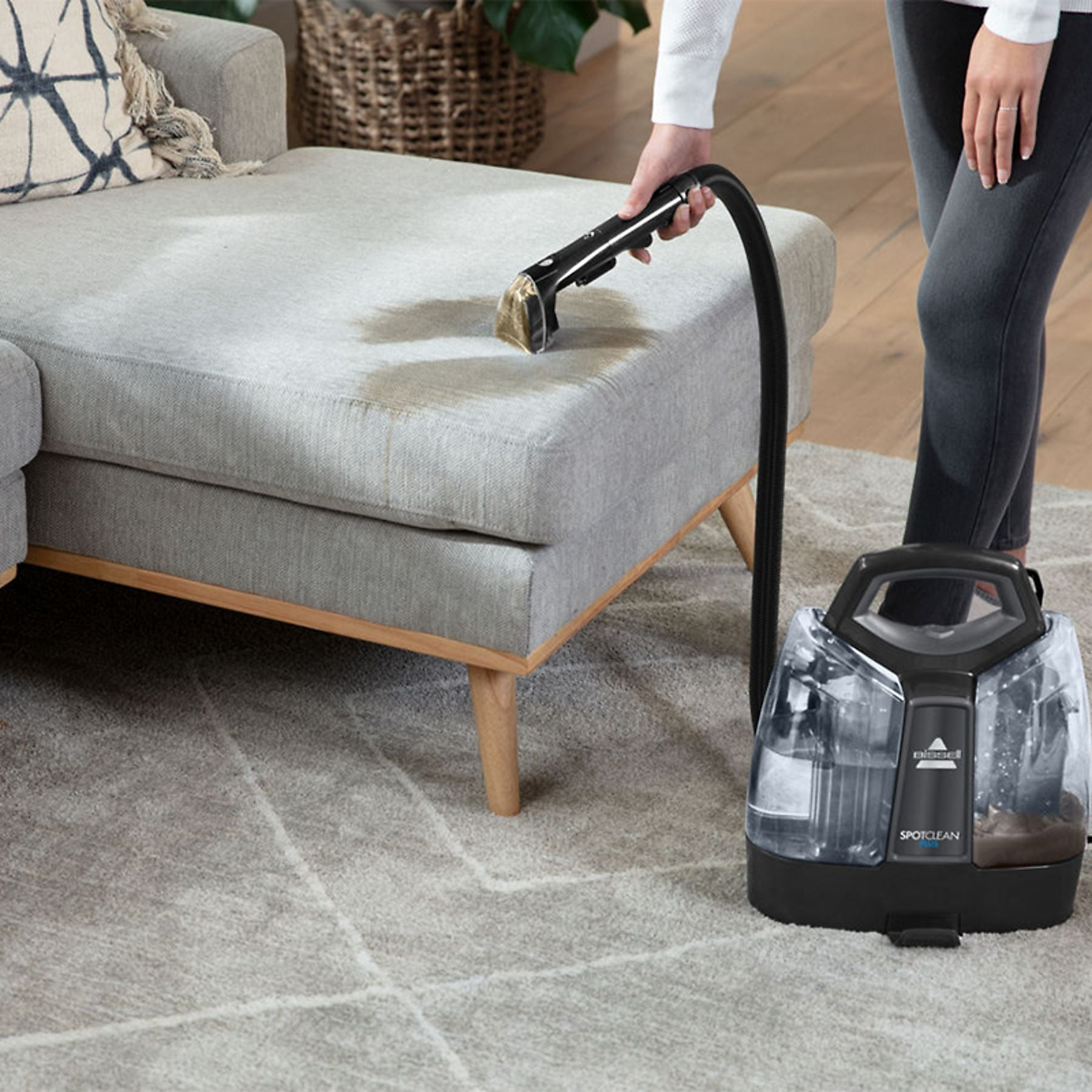 A person cleans a stain on a gray couch with a cleaning machine.