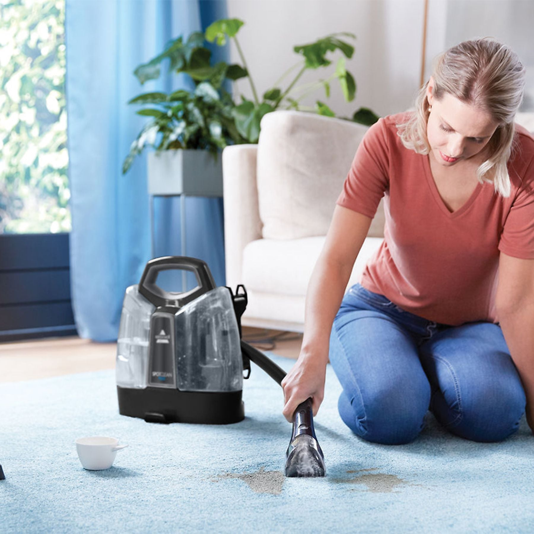 A woman kneels on a blue carpet using a spot cleaner on a stain.