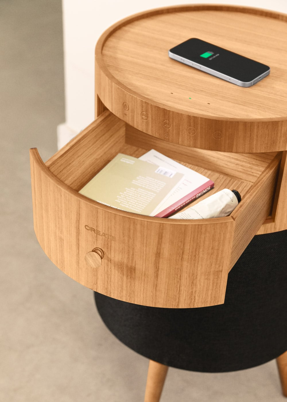 Mesita de noche de madera con cajón abierto, móvil encima, libro dentro.