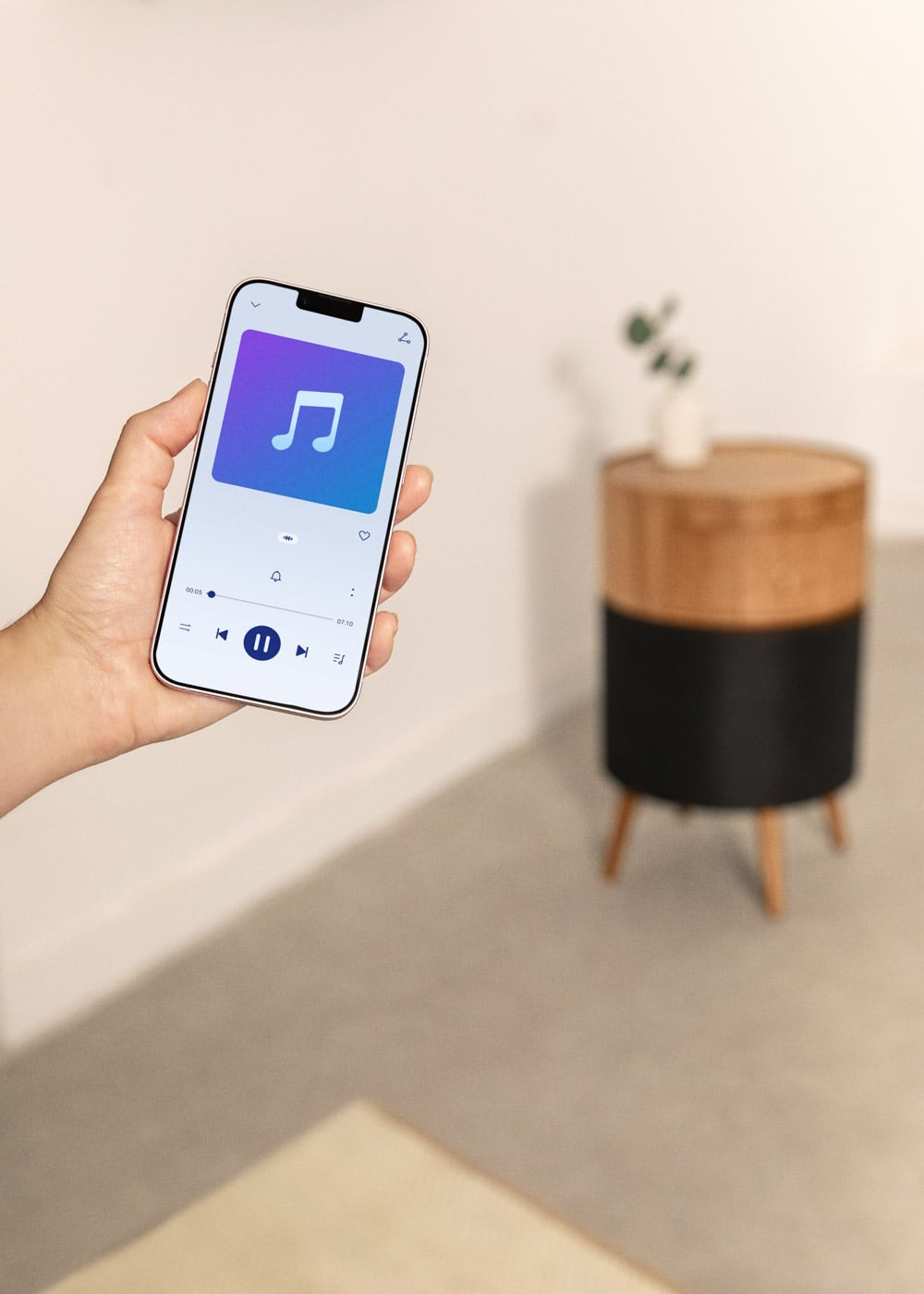 Mano sostiene smartphone con app de música, mesa auxiliar al fondo.
