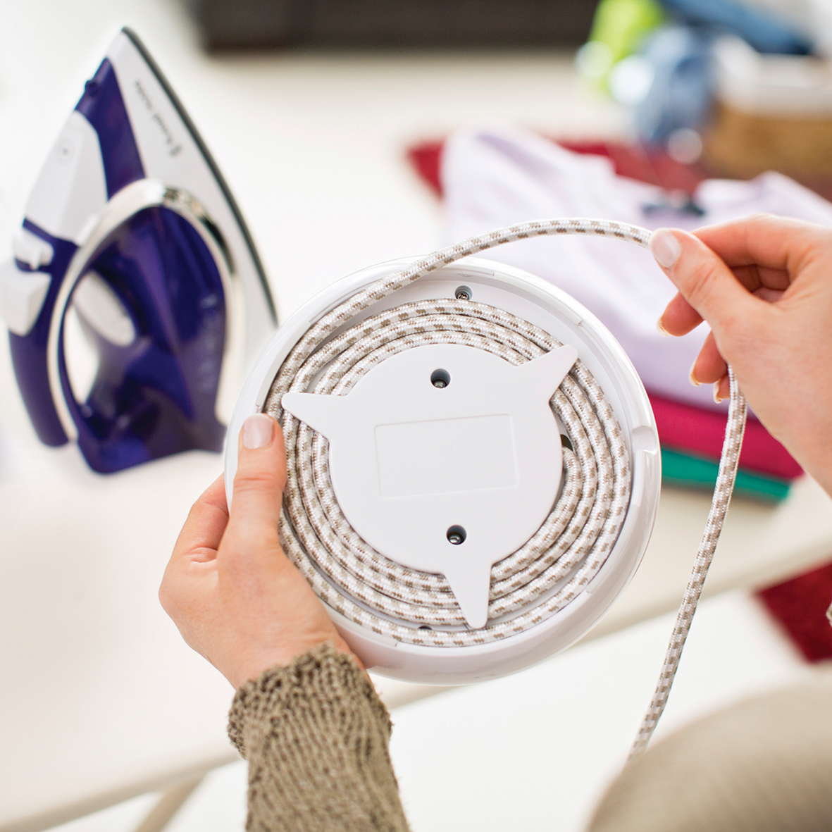 Osoba nawijająca sznur na szpulę; żelazko i ubrania w tle.