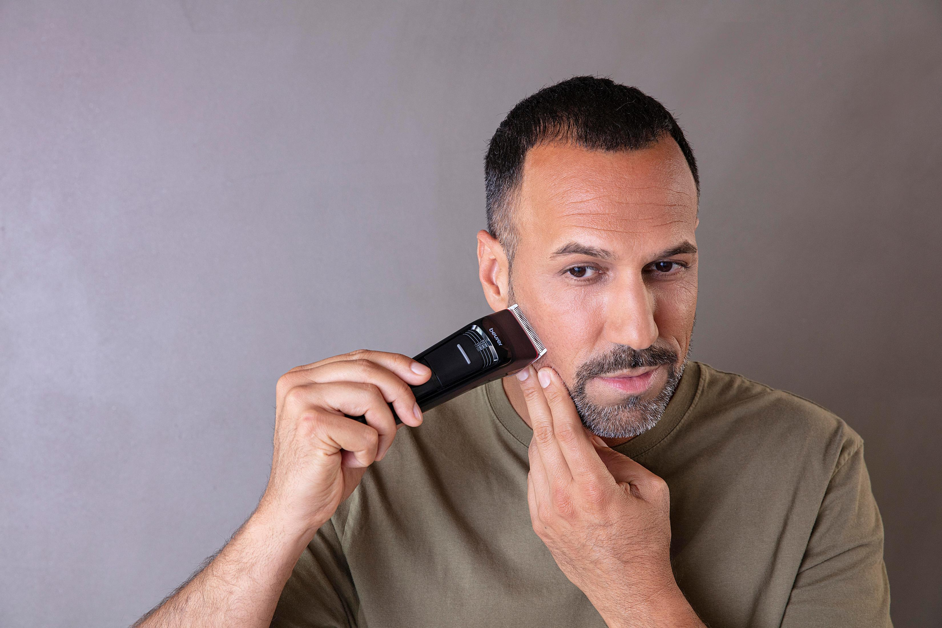 A man trims his beard with a black trimmer against a gray background.
