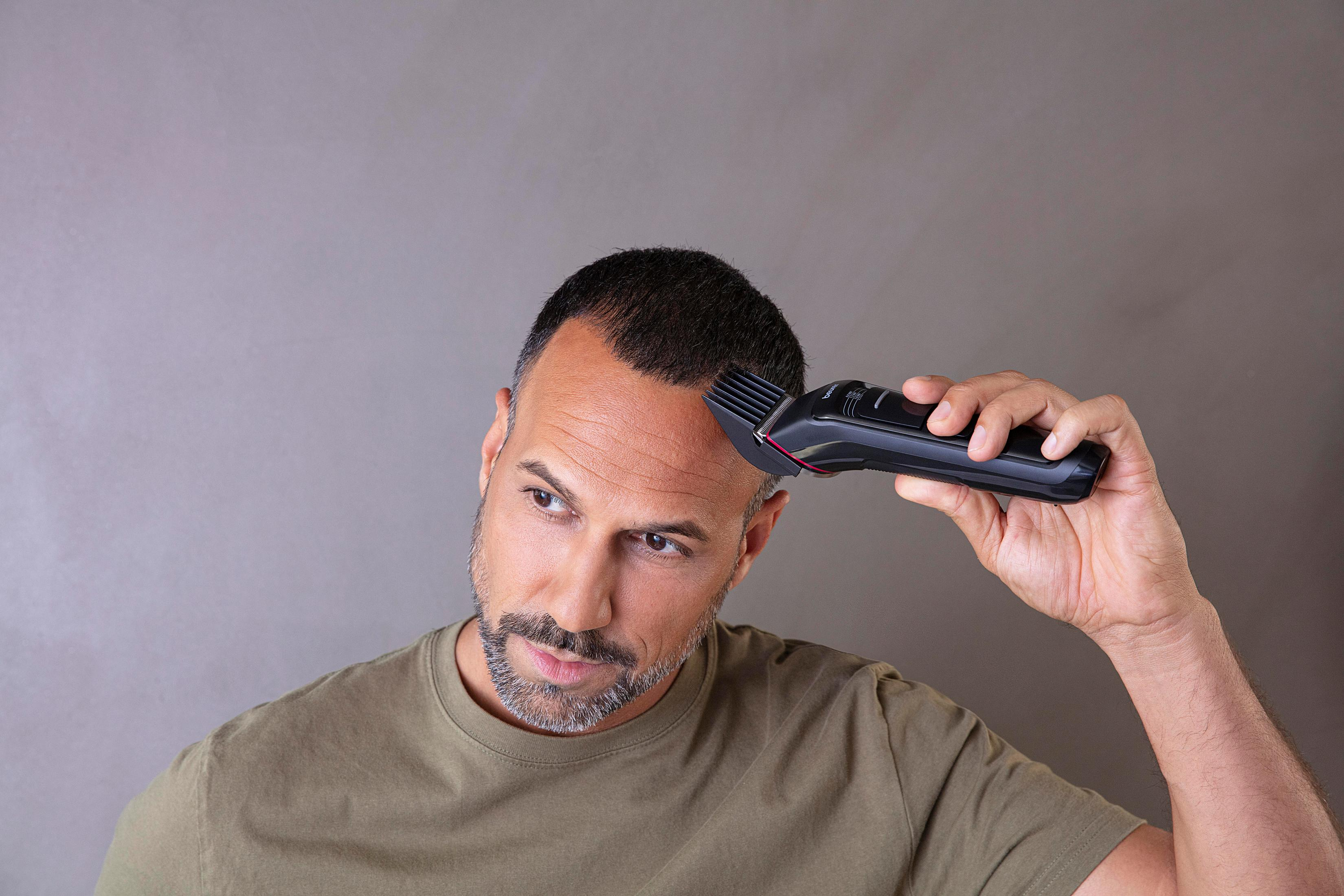 A man with a beard trims his hair with an electric hair clipper on his head.