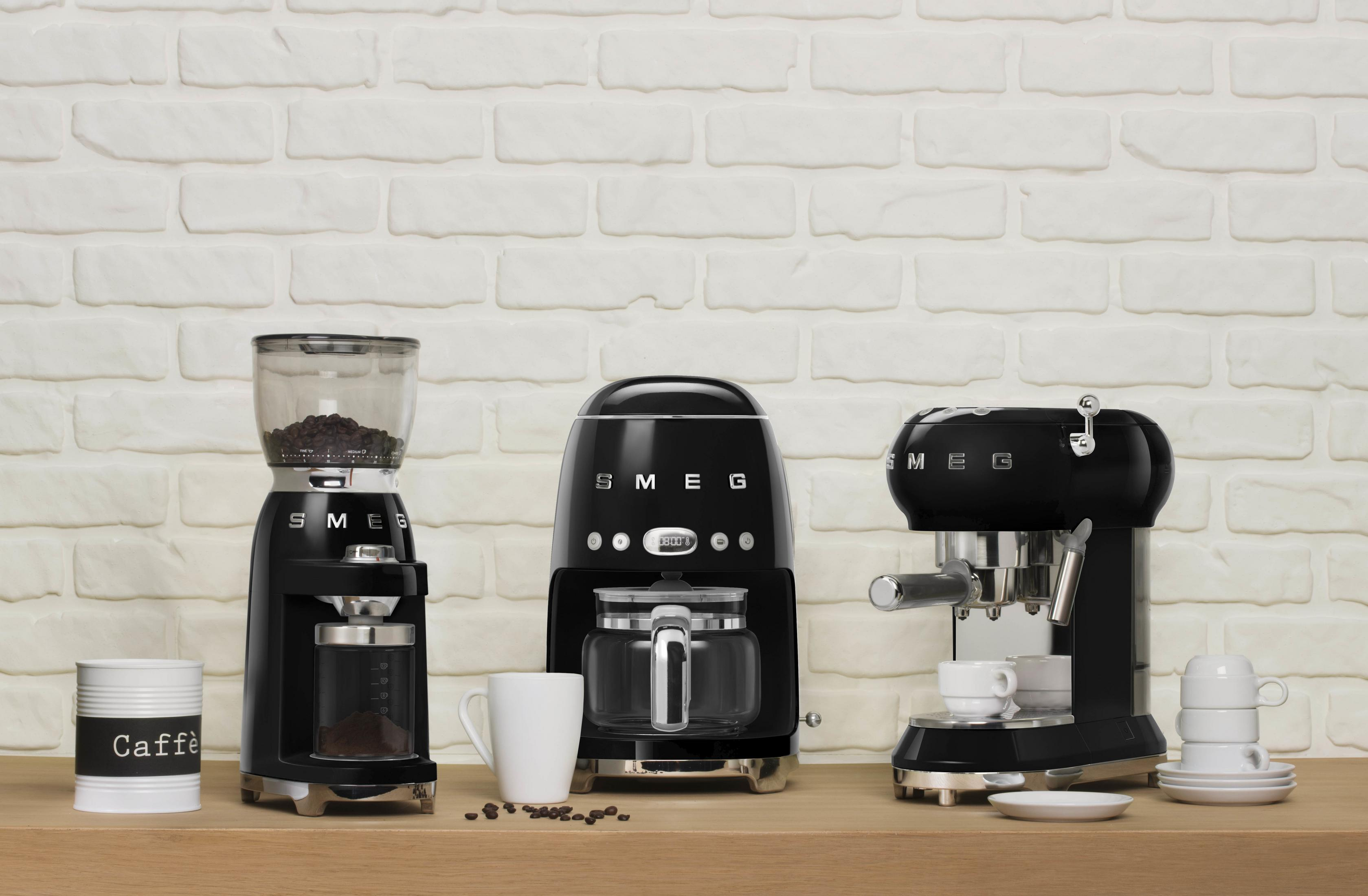 Trzy czarne ekspresy do kawy SMEG stoją na drewnianym stole.