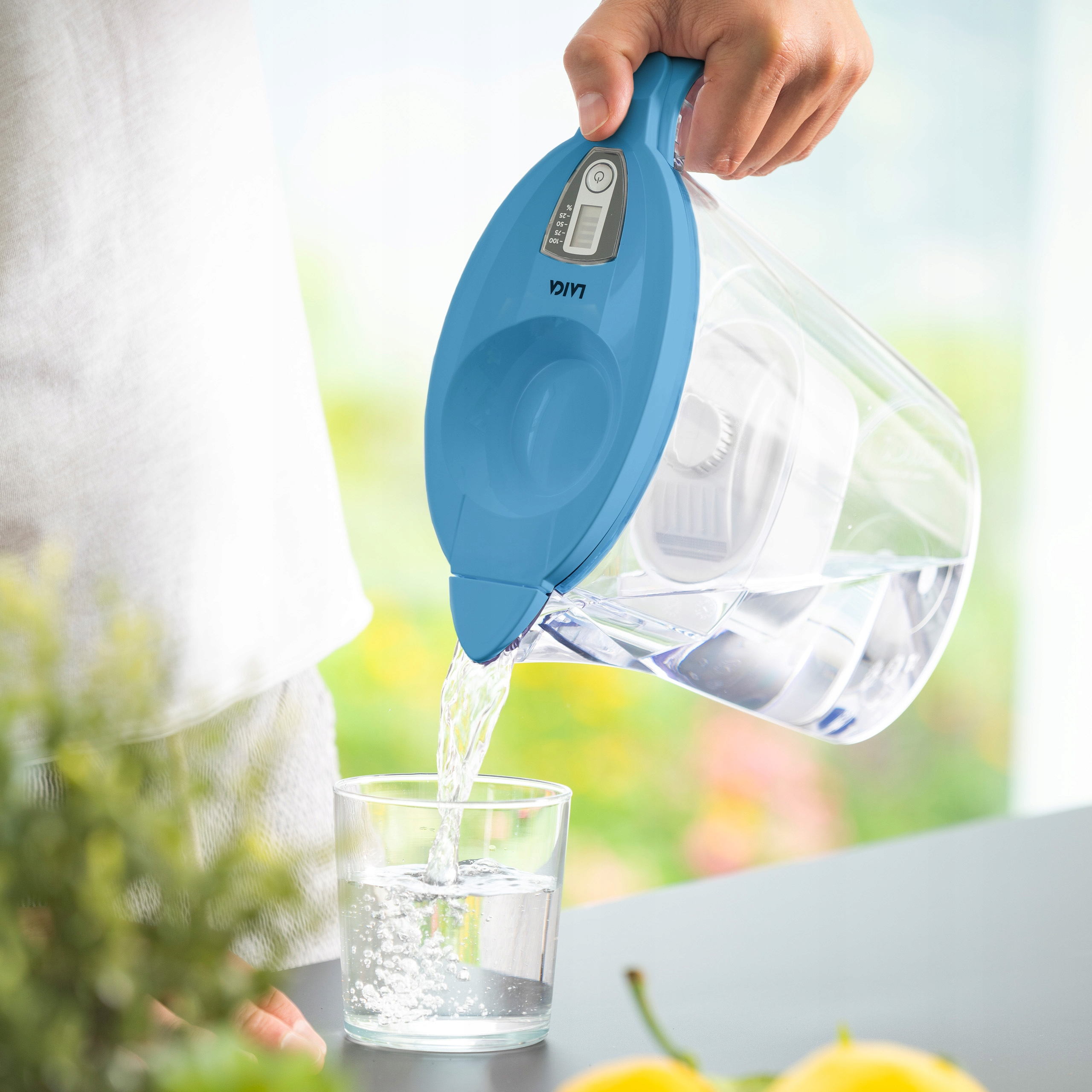 Ręka przelewa wodę z niebieskiego dzbanka do szklanki. Rozmyty zielony krajobraz w tle.