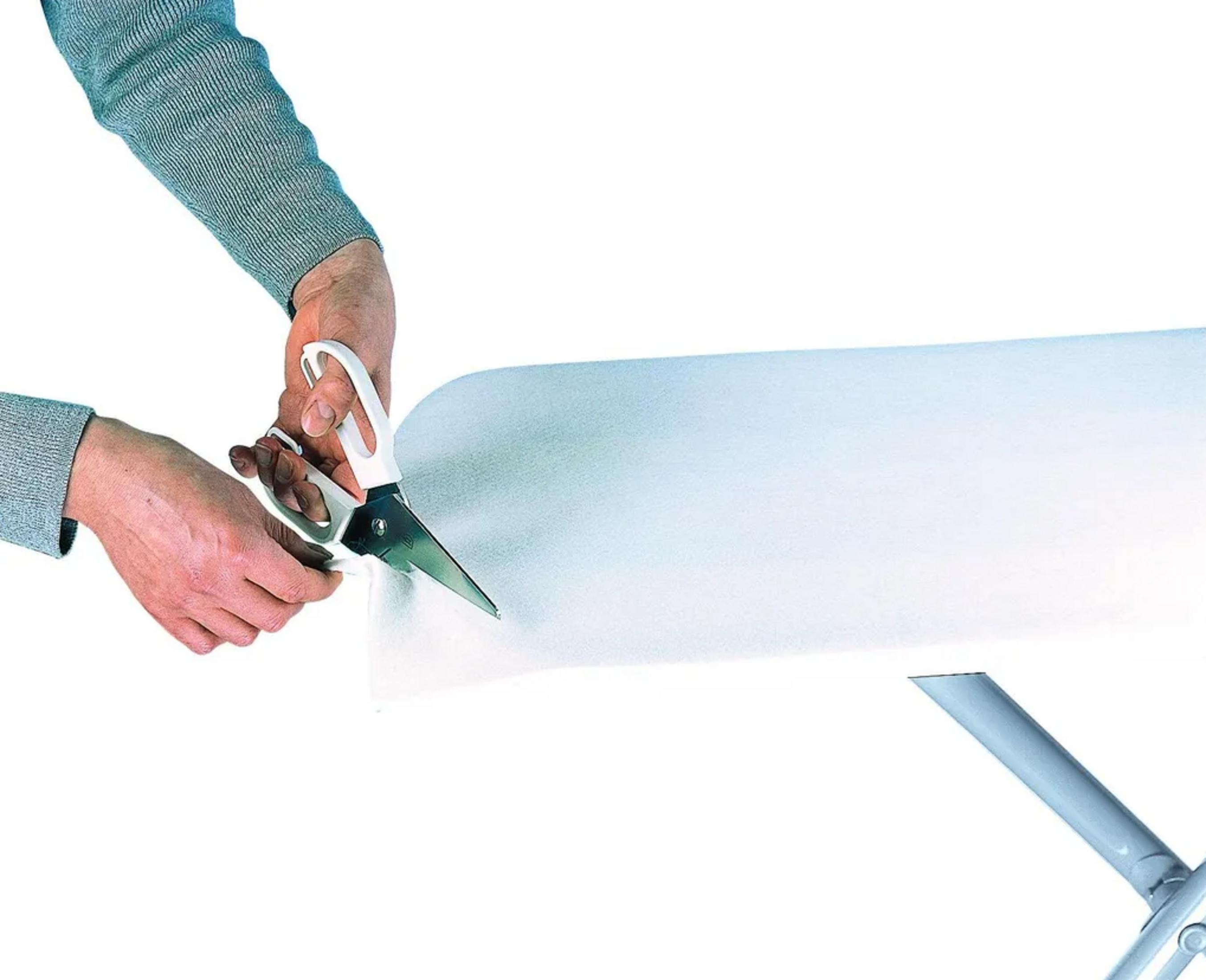 Dłonie przecinające biały materiał nożyczkami nad ramą deski do prasowania.