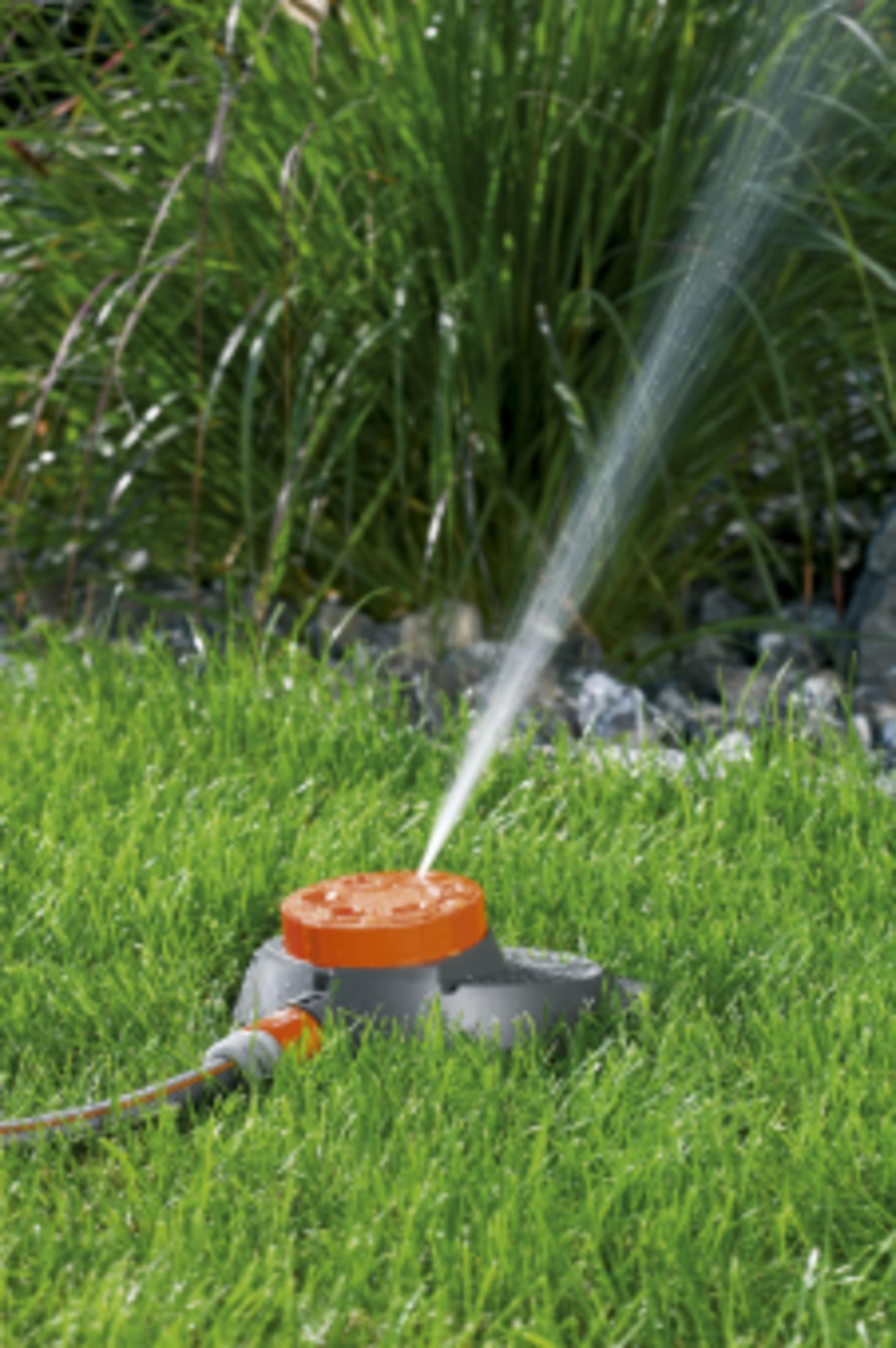 Ein Gartensprenger mit orangem Oberteil bewässert grünes Gras und Pflanzen.