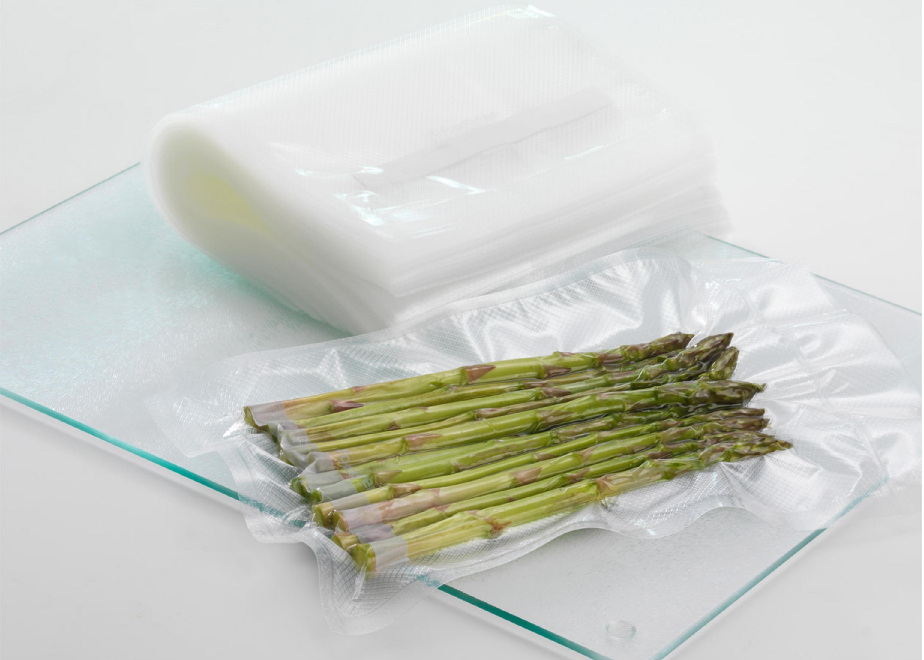 Grüner Spargel in Vakuumbeutel auf Glasplatte, Stapel Beutel daneben.