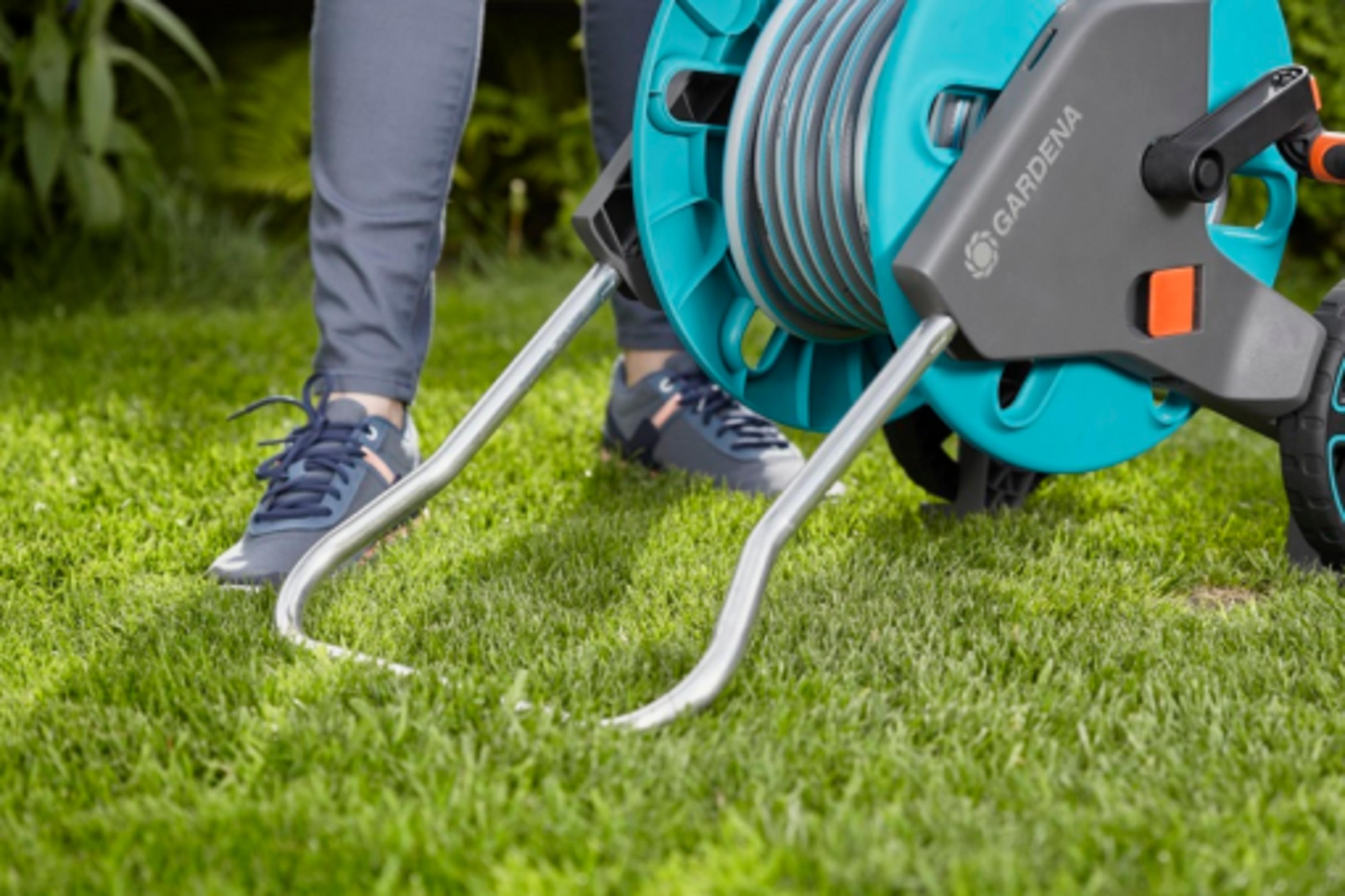 Eine Person steht im Gras neben einem Gartenschlauchwagen mit blauem und grauem Gehäuse.