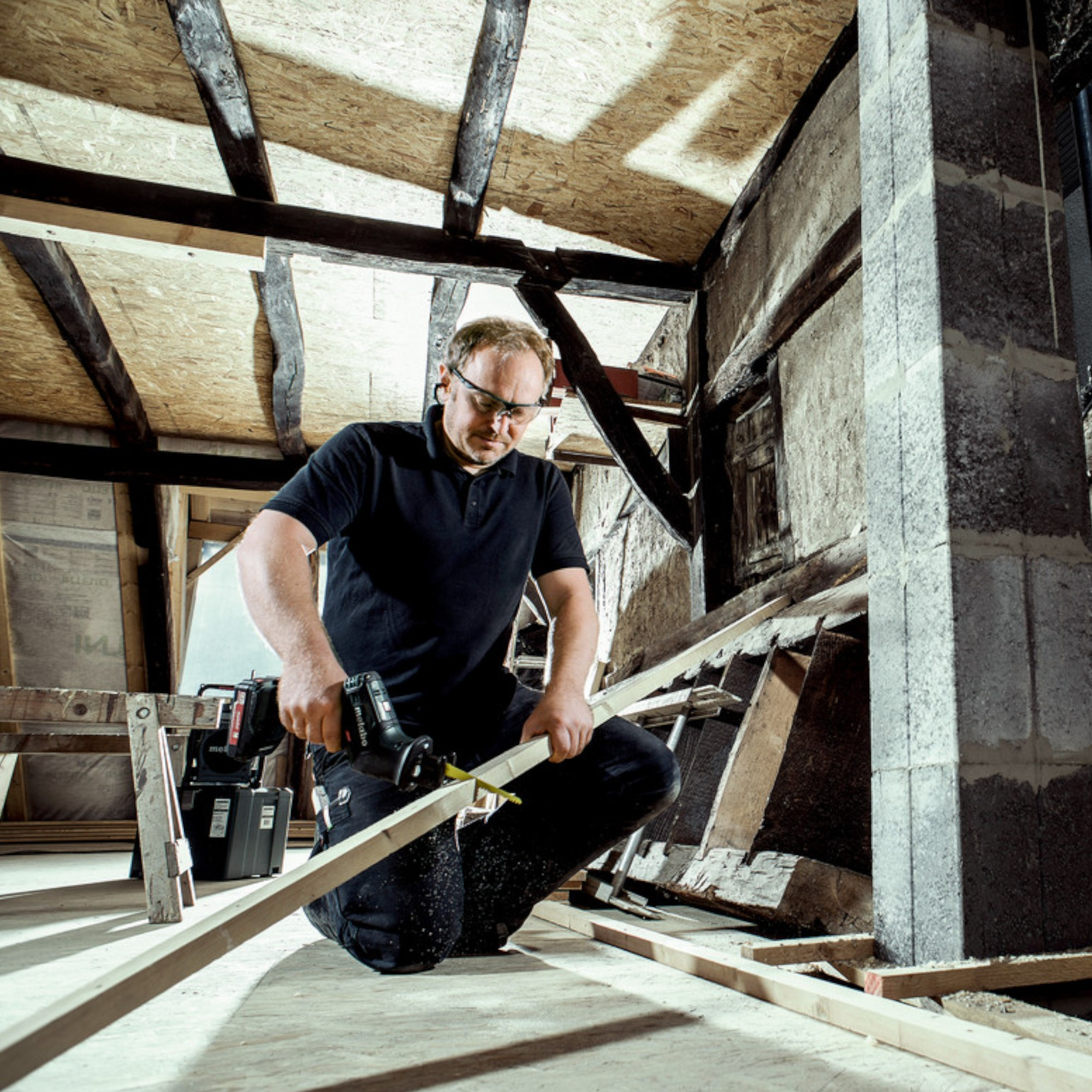 Ein Mann in schwarzer Kleidung sägt Holz in einem Rohbau.
