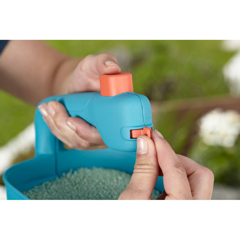 Eine Hand betätigt einen orangefarbenen Knopf an einem blauen Streuer mit Granulat.