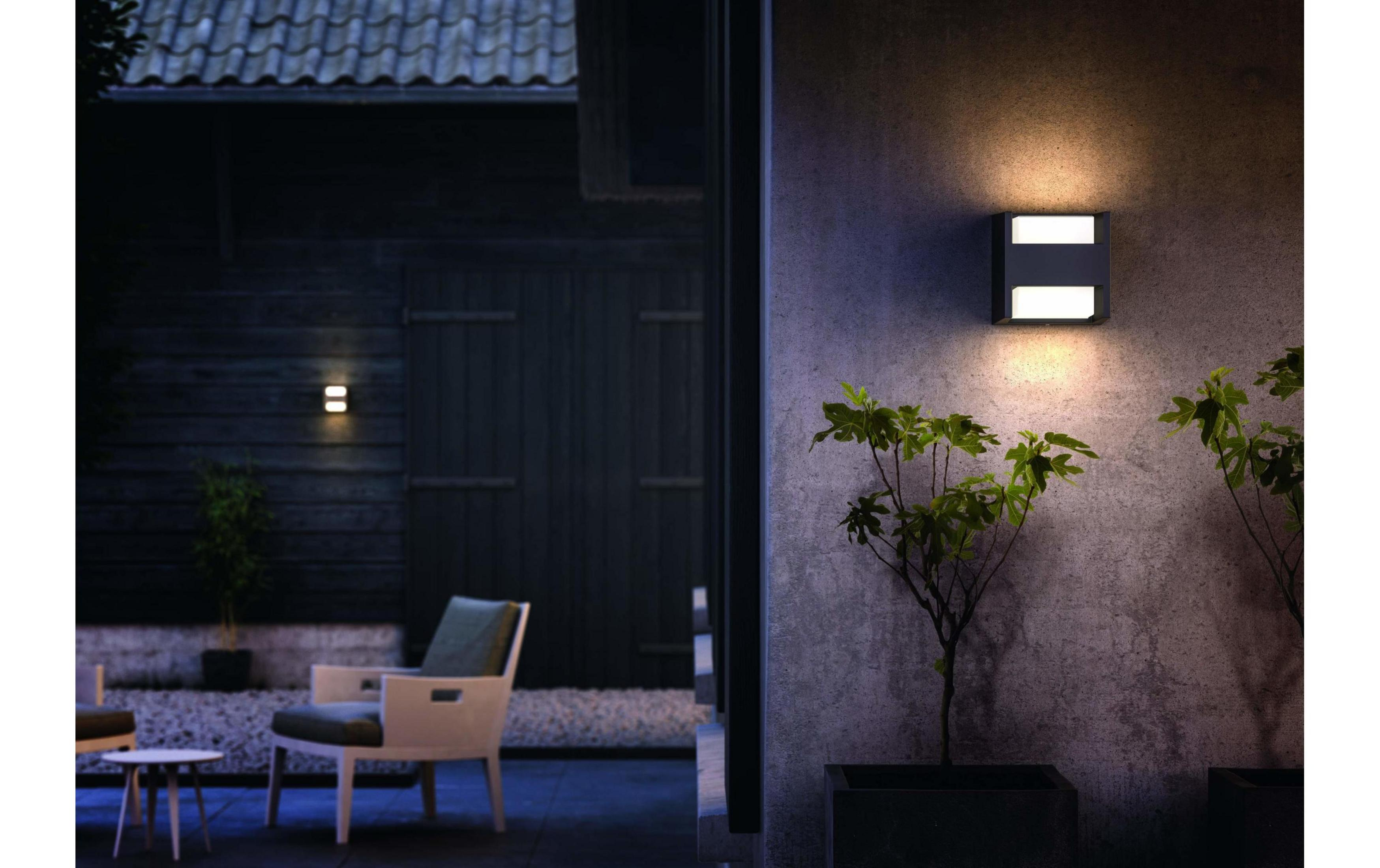 Lámpara de pared exterior ilumina muro de hormigón y plantas de noche.