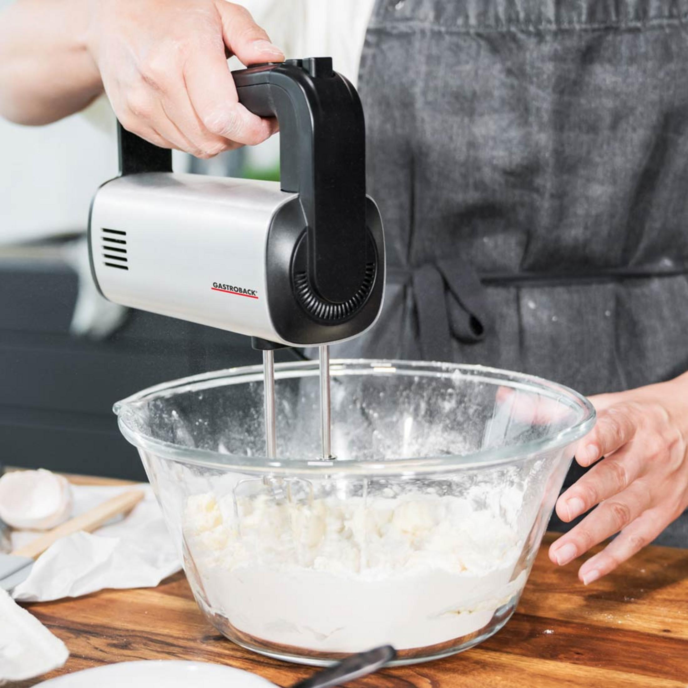 Una persona usa uno sbattitore elettrico a mano d'argento per mescolare ingredienti in una ciotola di vetro.