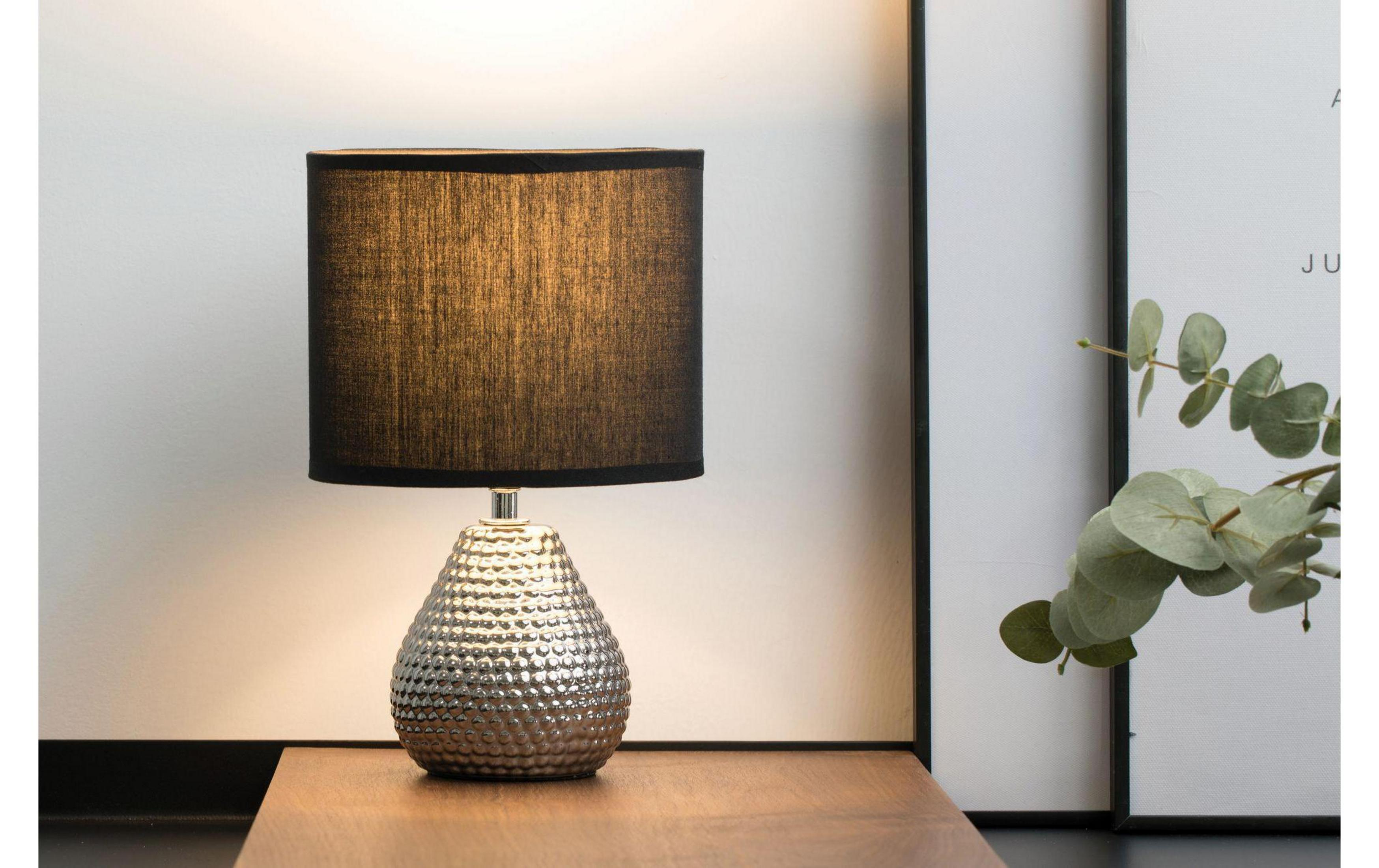 Srebrna lampa z czarnym kloszem na drewnianym stole, obok obrazu i eukaliptusa.