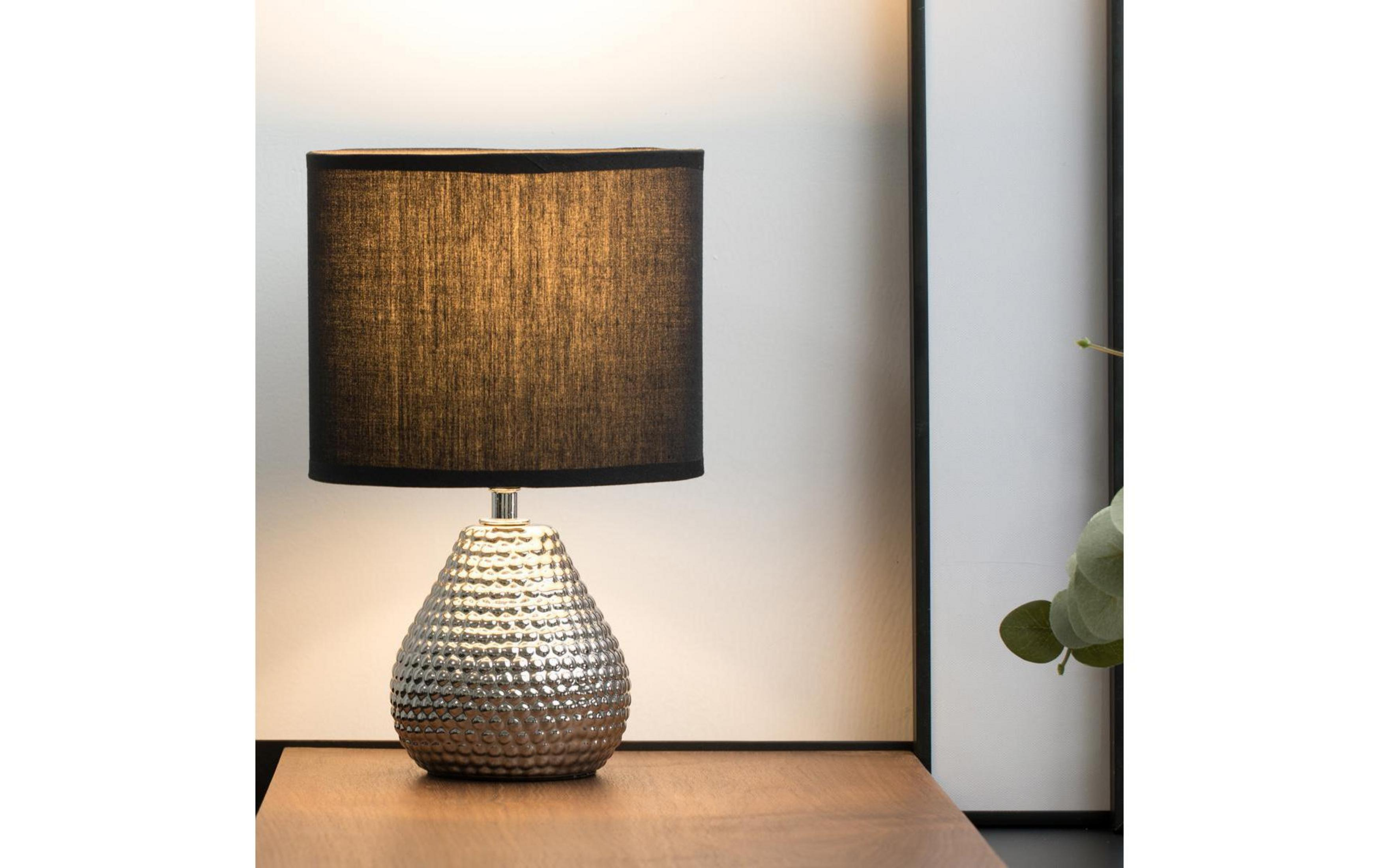 Srebrna lampa z teksturowaną podstawą i czarnym kloszem stoi na drewnianym stole.