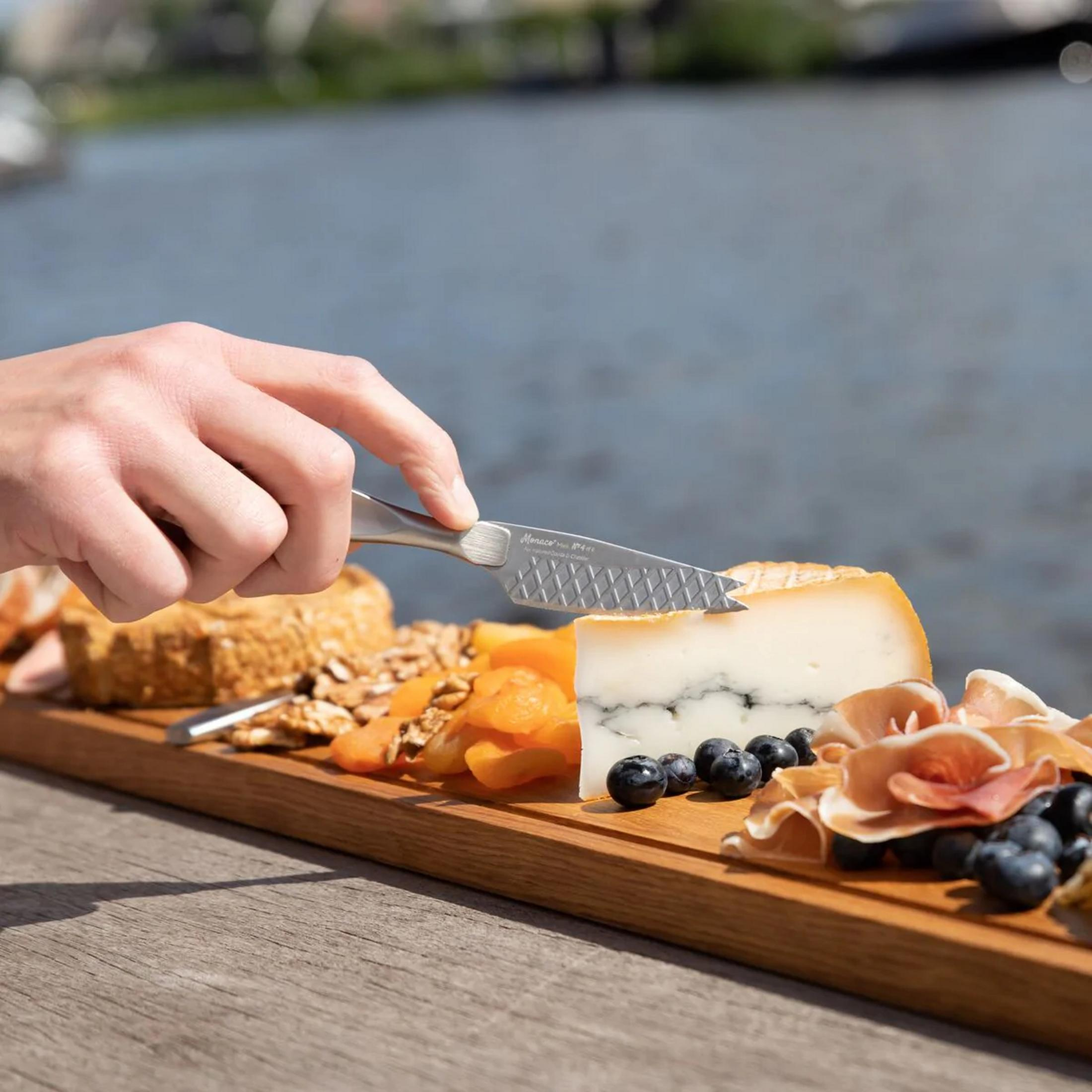 A hand uses a cheese knife on a charcuterie board with cheese, nuts, dried apricots, blueberries, and ham.