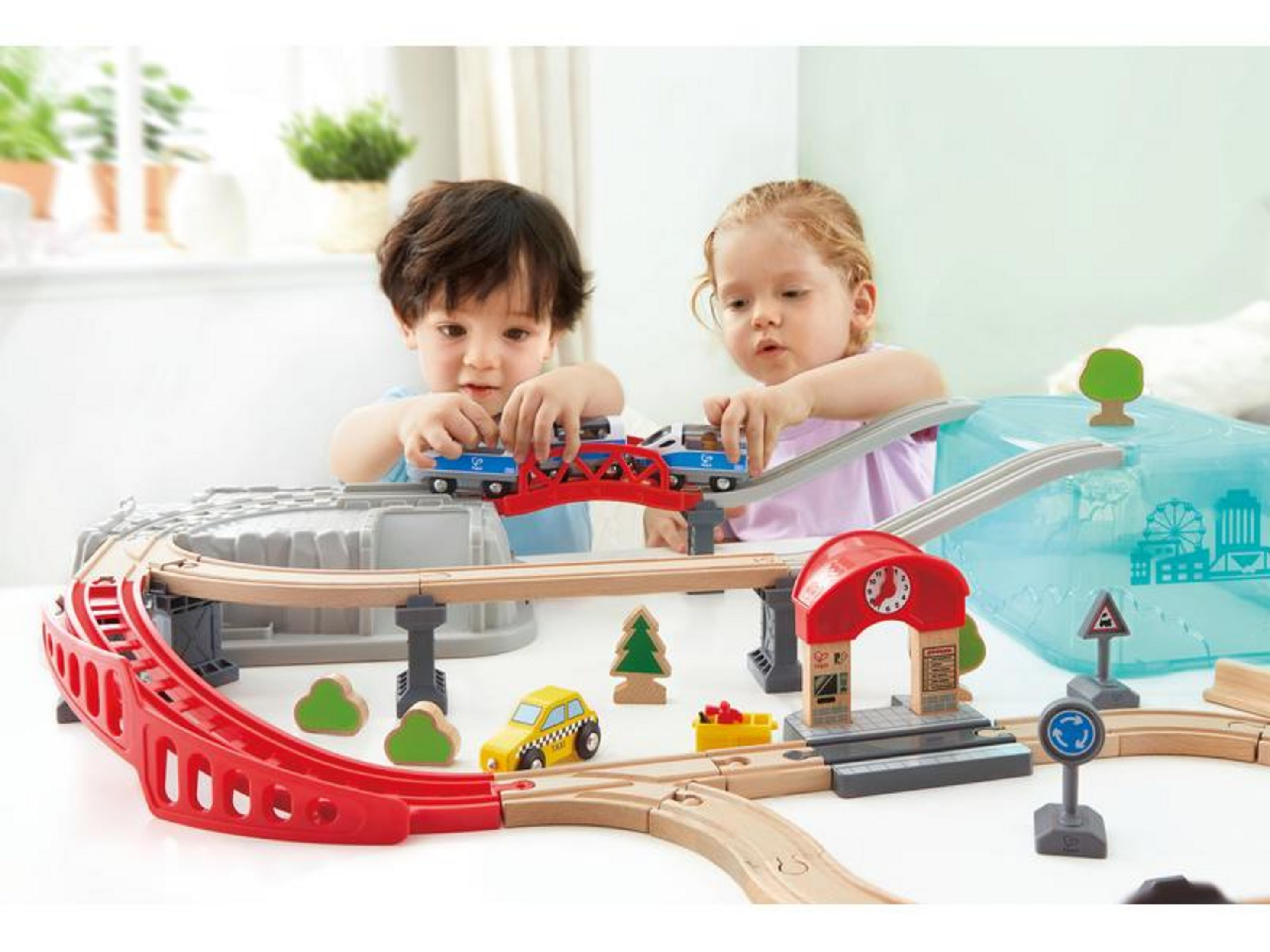 Dzieci bawią się drewnianą kolejką, z pociągami, samochodzikami i budynkami.