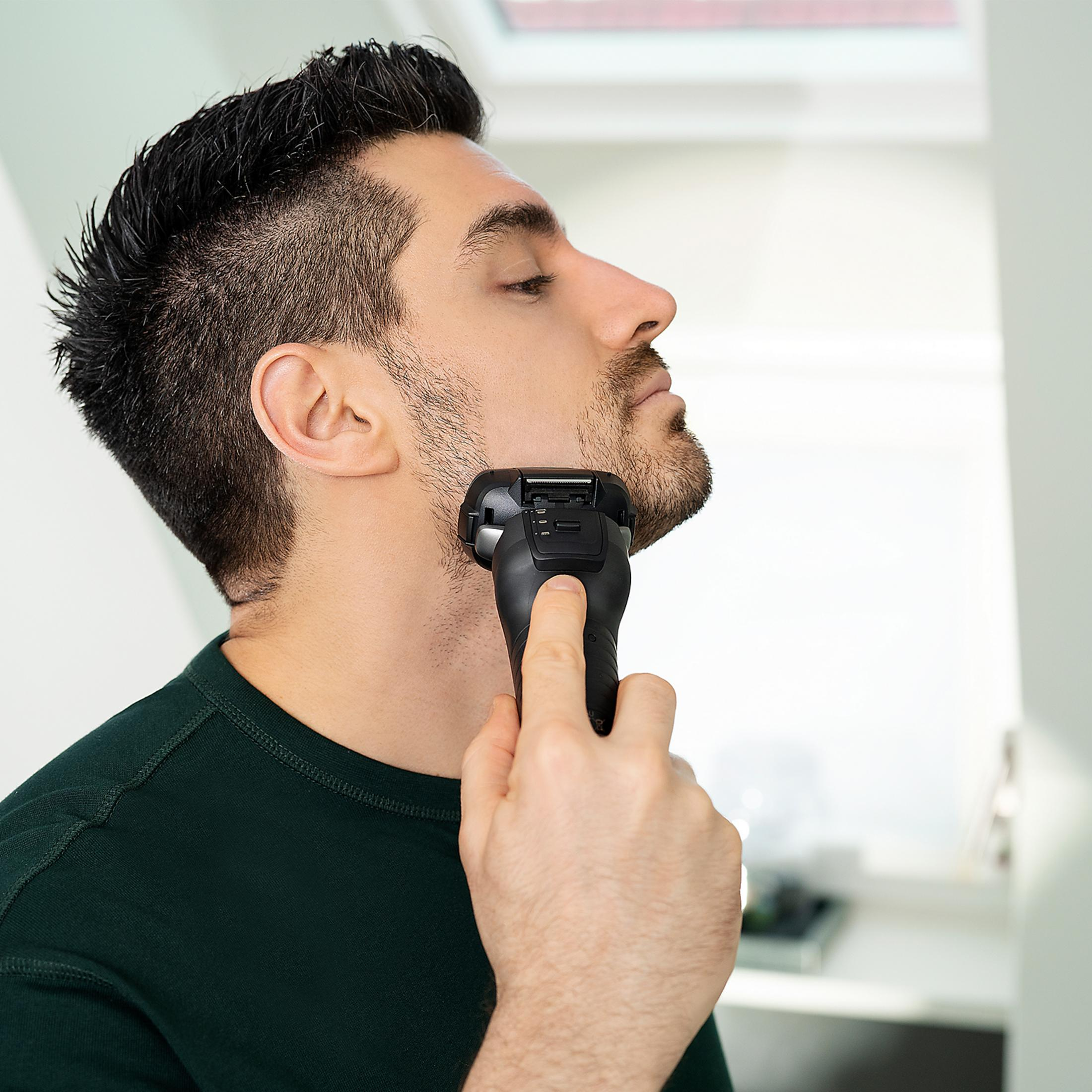 Mężczyzna goli twarz golarką elektryczną. Nosi ciemnozieloną koszulkę.