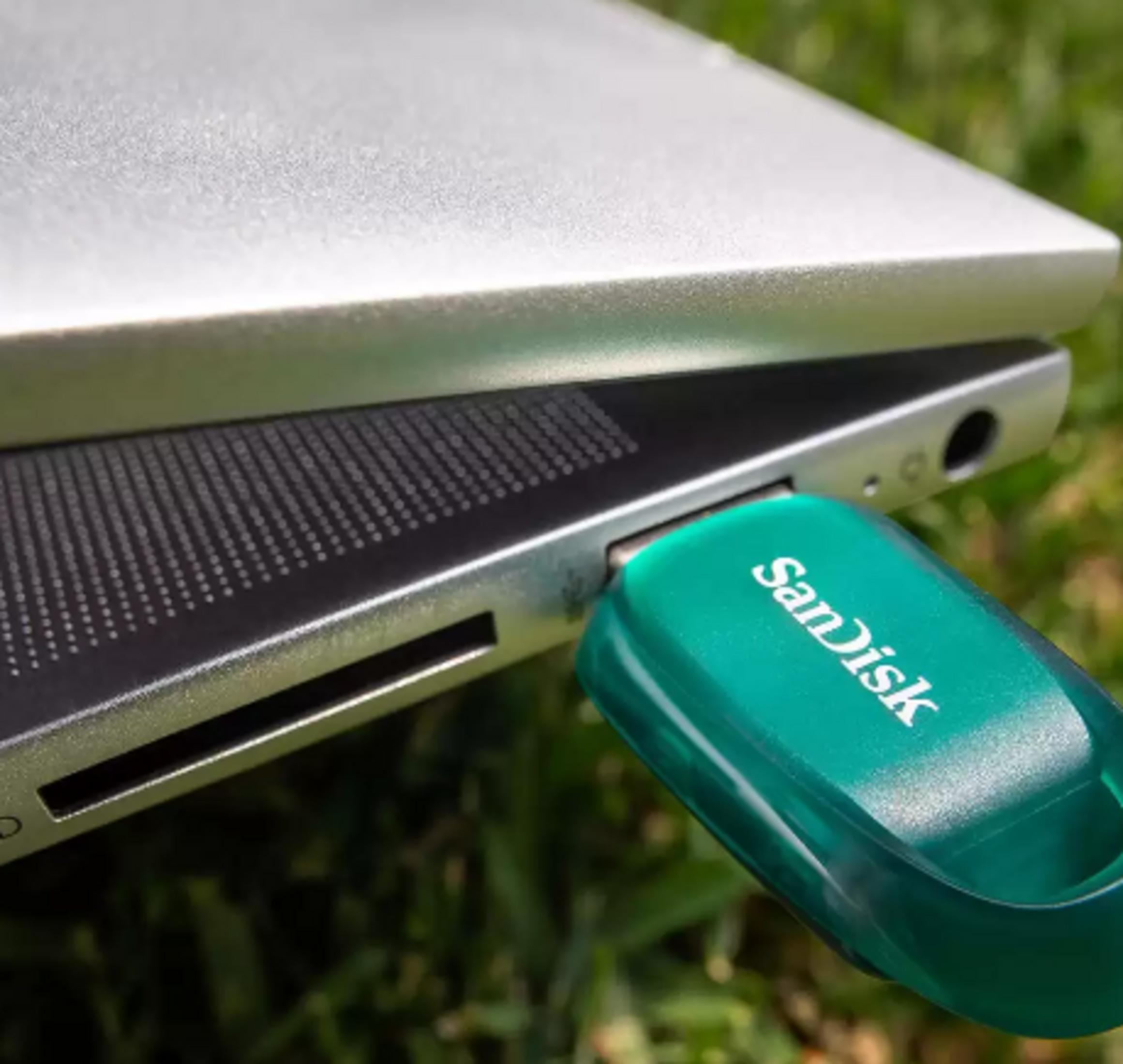 Zielony pendrive SanDisk podłączony do srebrnego laptopa, na zewnątrz.