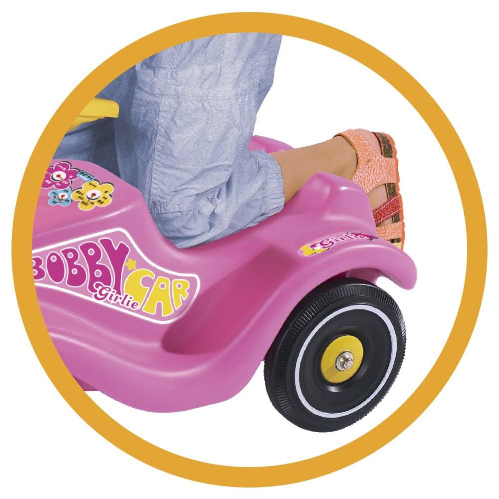 Ein Kind in blauer Jeans und Sandalen sitzt auf einem pinken Bobby Car mit 'Girlie'-Aufkleber.