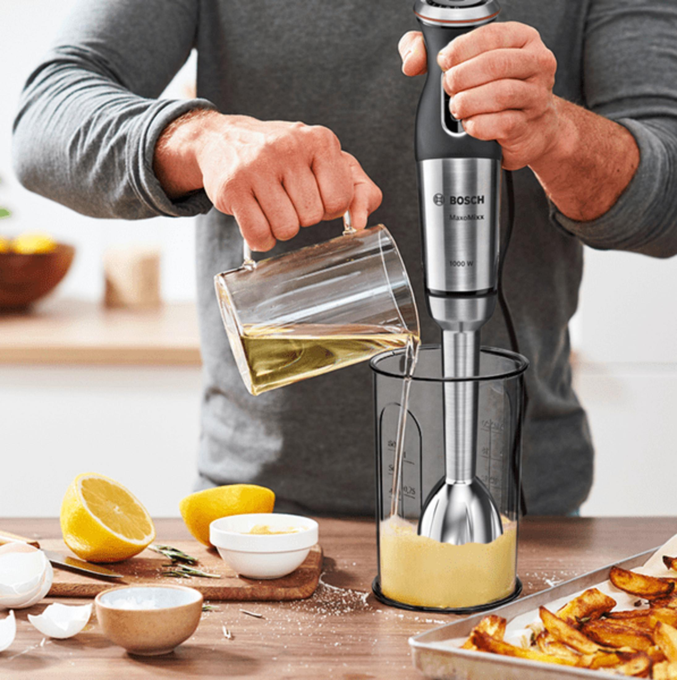 Mann gießt Öl in einen Mixerbecher, daneben Zitronen und Kartoffelspalten.