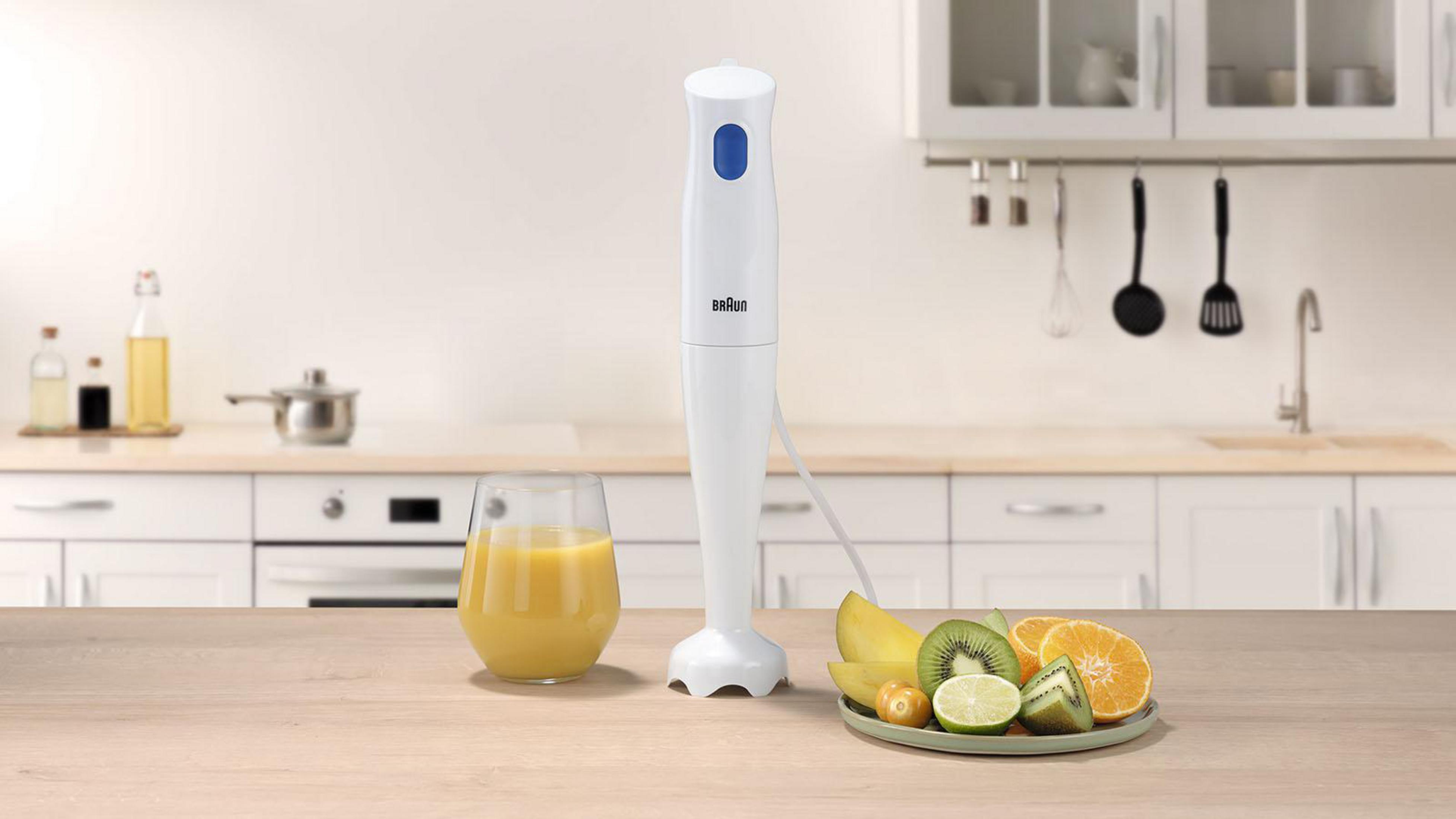 Biały blender ręczny stoi obok szklanki soku pomarańczowego i miski z owocami.