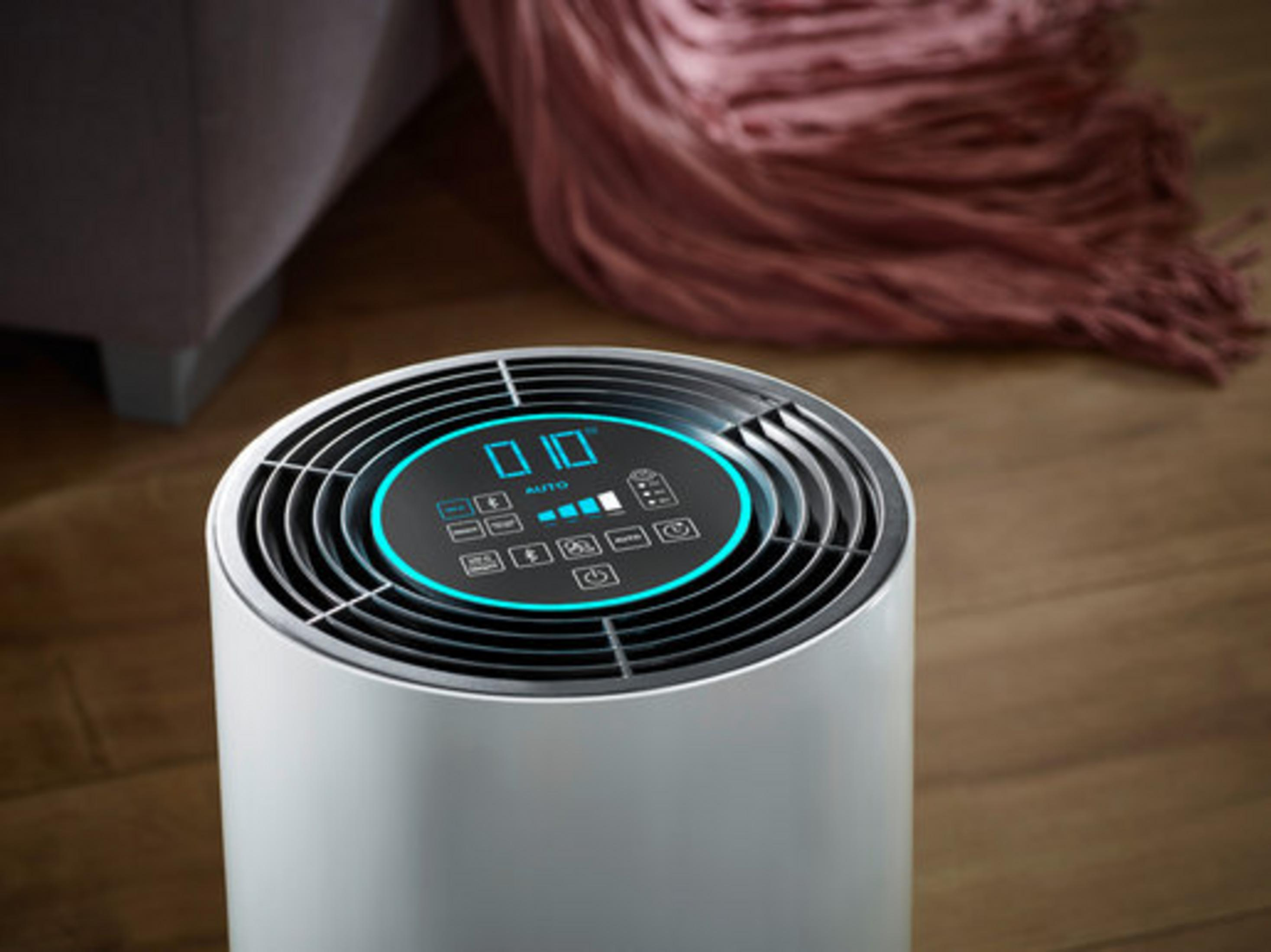 Ein weißer Luftreiniger mit blau beleuchtetem Display steht auf einem Holzboden.