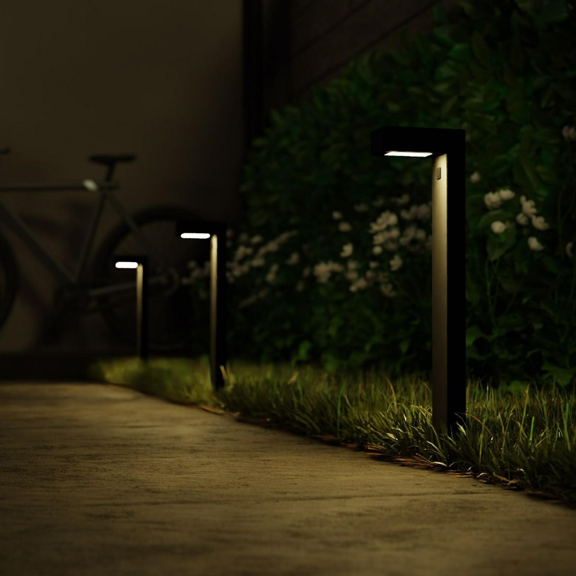 Modernas luces de camino iluminan un sendero junto a césped y arbustos. Una bicicleta es visible al fondo.