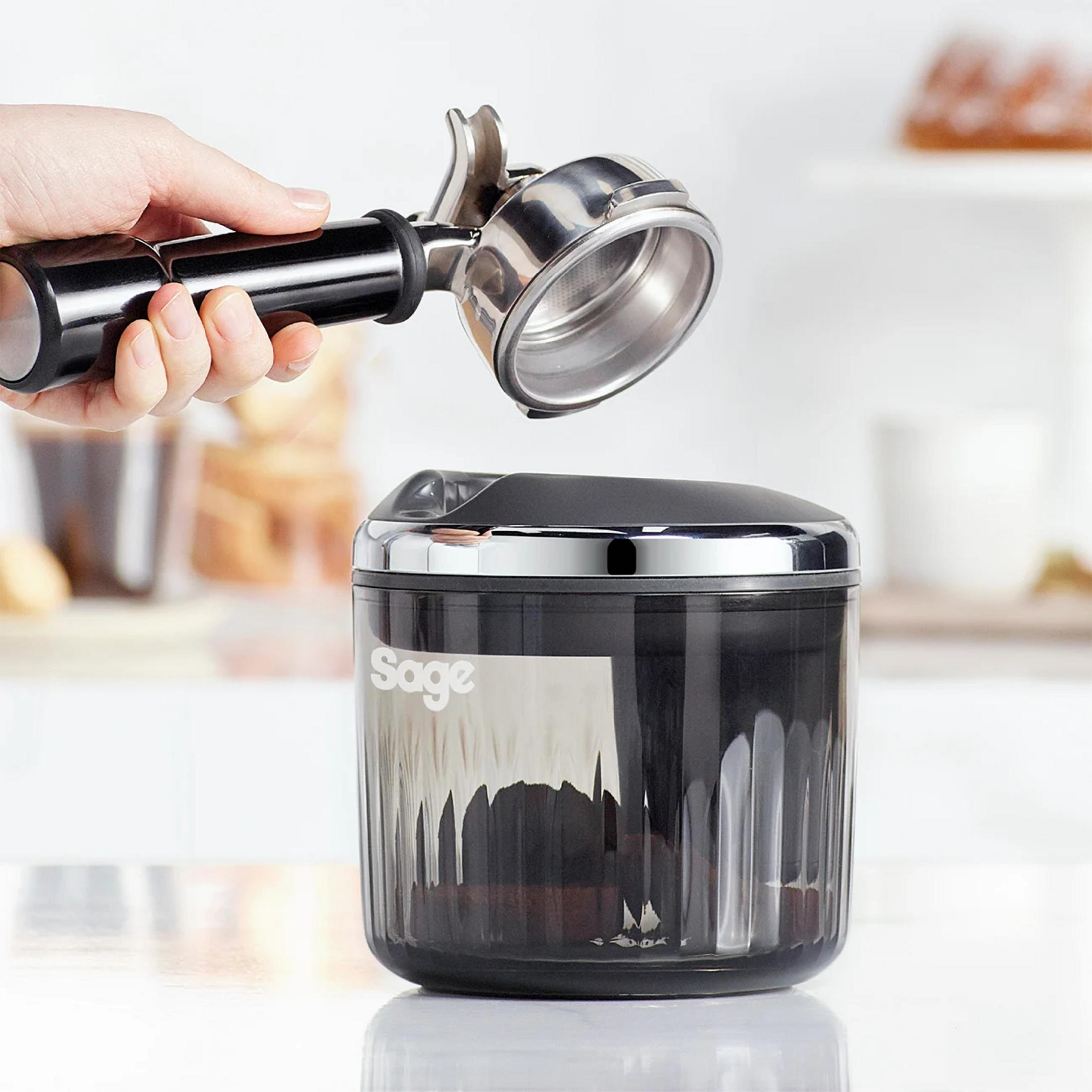 Una mano tiene un portafiltro sopra un contenitore nero per fondi di caffè Sage.