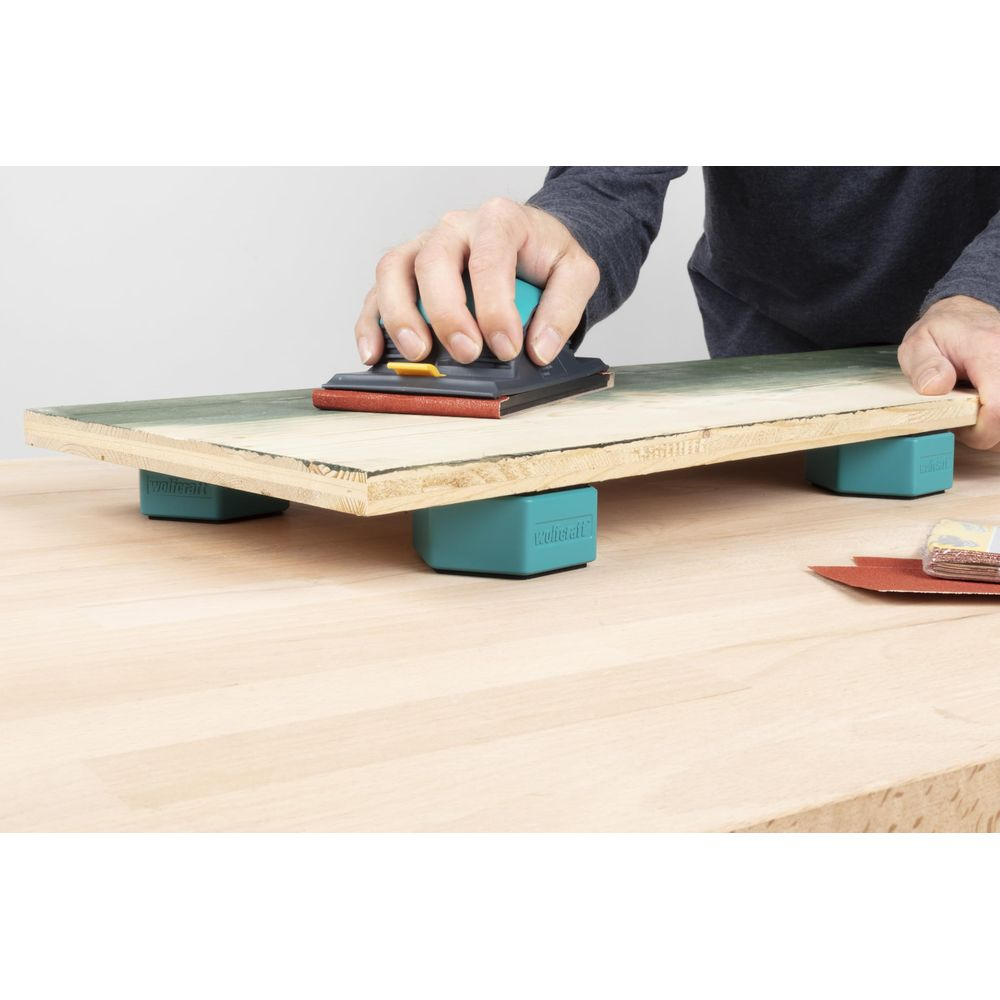 Main ponçant une planche en bois sur des blocs turquoise. Papier de verre à côté.