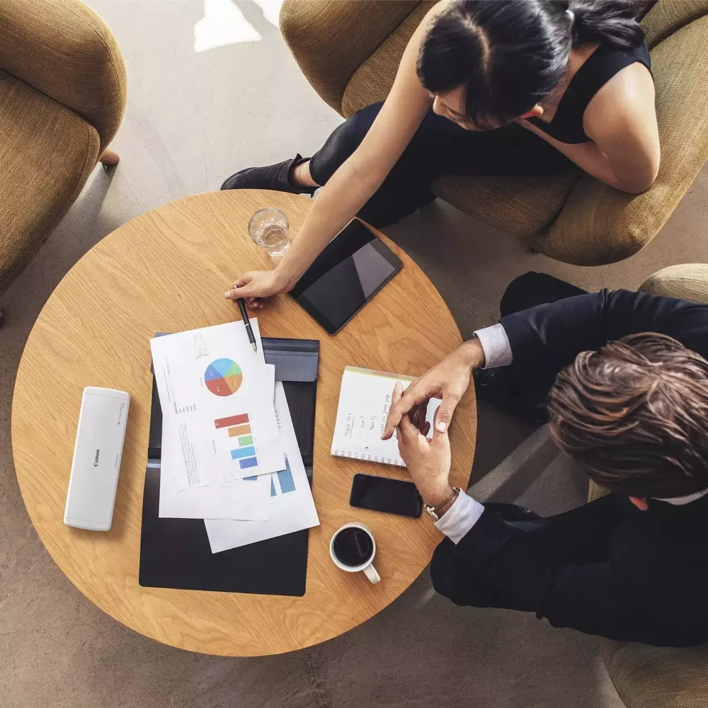Zwei Personen diskutieren über Diagramme an einem Holztisch mit Kaffee und Tablet.