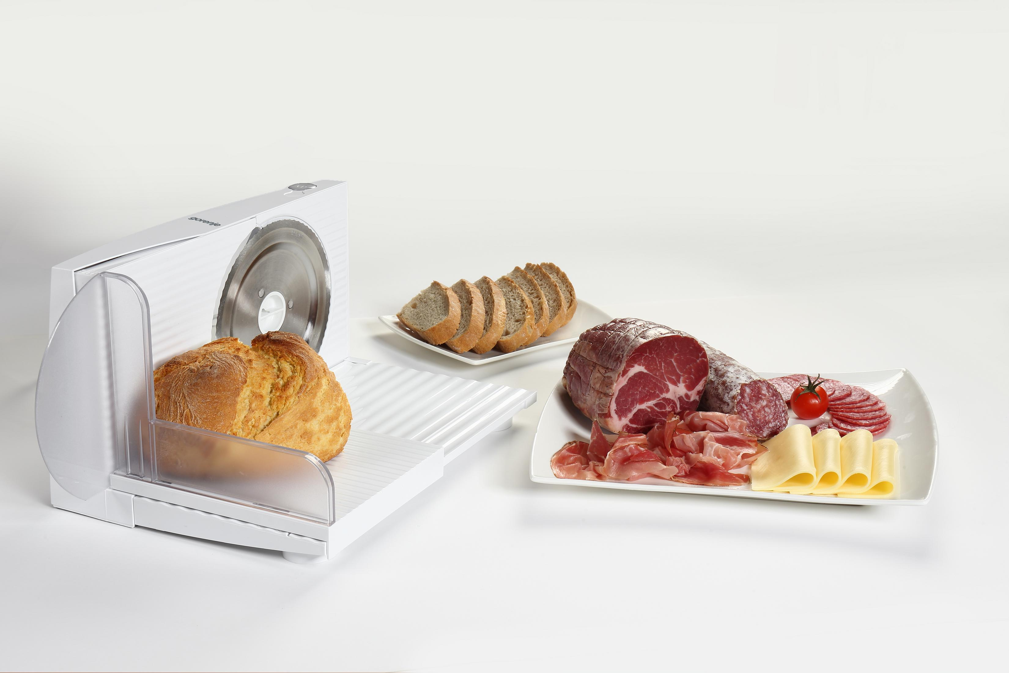 Krajalnica z chlebem, salami, szynką, serem i pomidorem na talerzu.