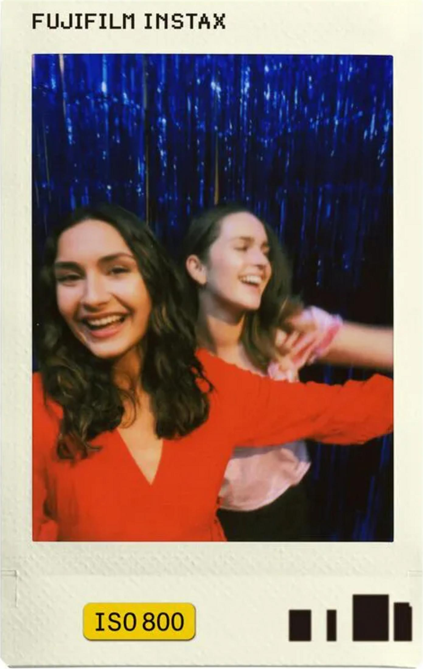 Dos mujeres jóvenes ríen ante una cortina de lentejuelas azules. La de delante lleva rojo.