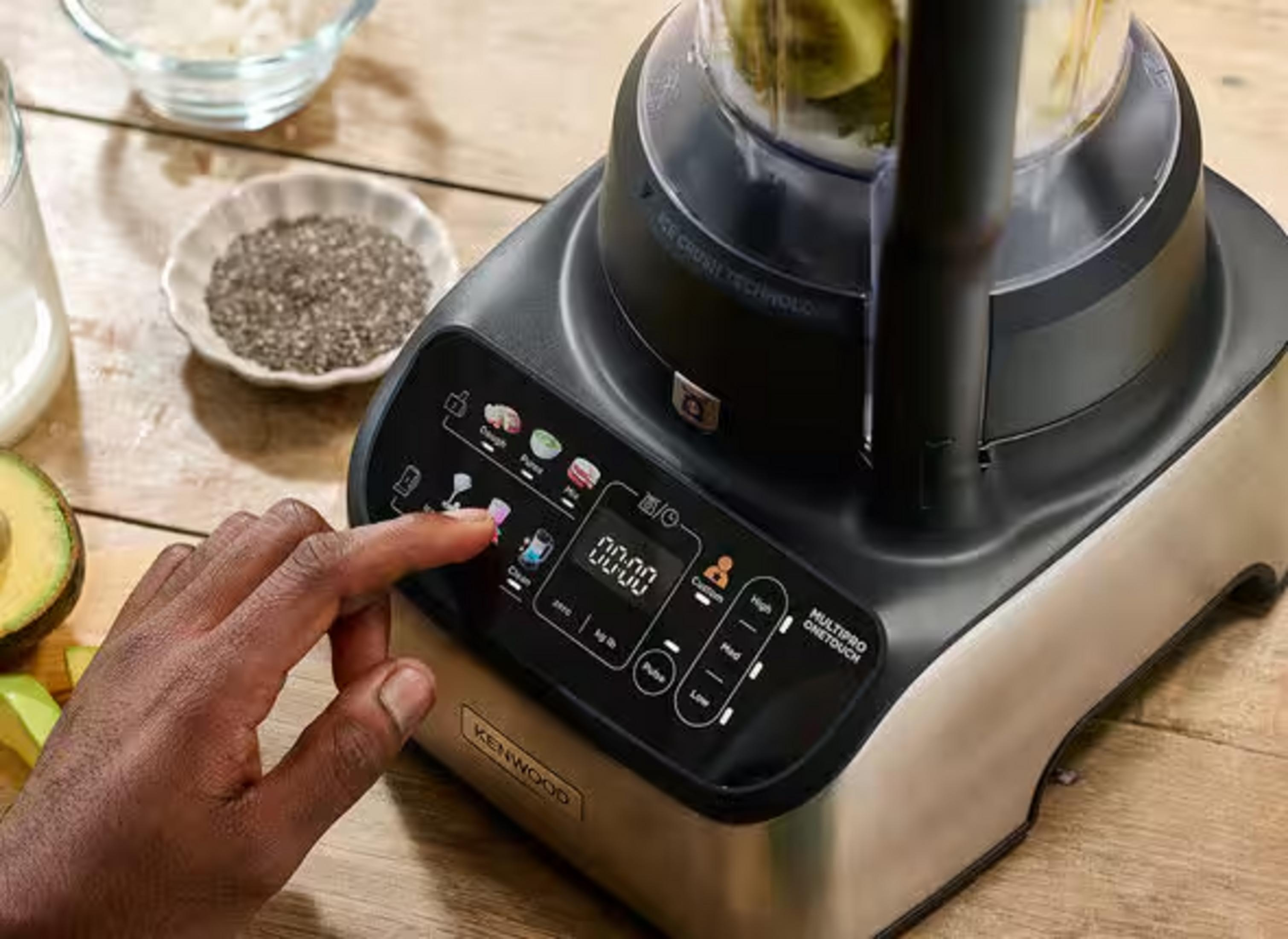 Una mano aziona il pannello di controllo di un robot da cucina Kenwood con avocado e semi di chia.