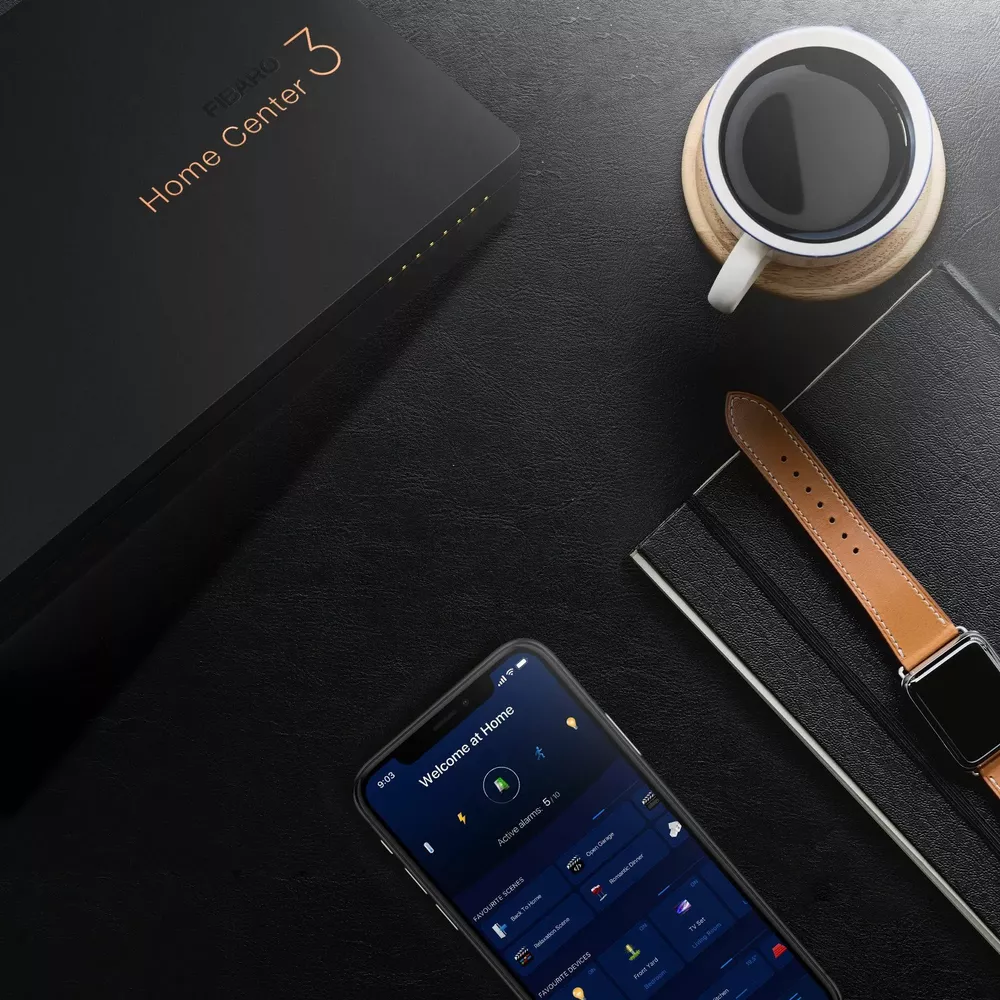 Ein schwarzes Fibaro Home Center 3 Gerät, ein Smartphone mit App, eine Tasse Kaffee und eine Uhr.