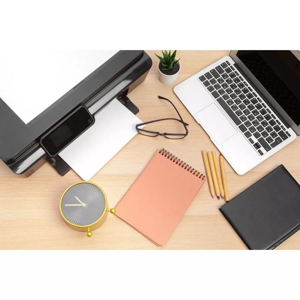 Biurko z drukarką, laptopem, notatnikiem, zegarem i ołówkami.