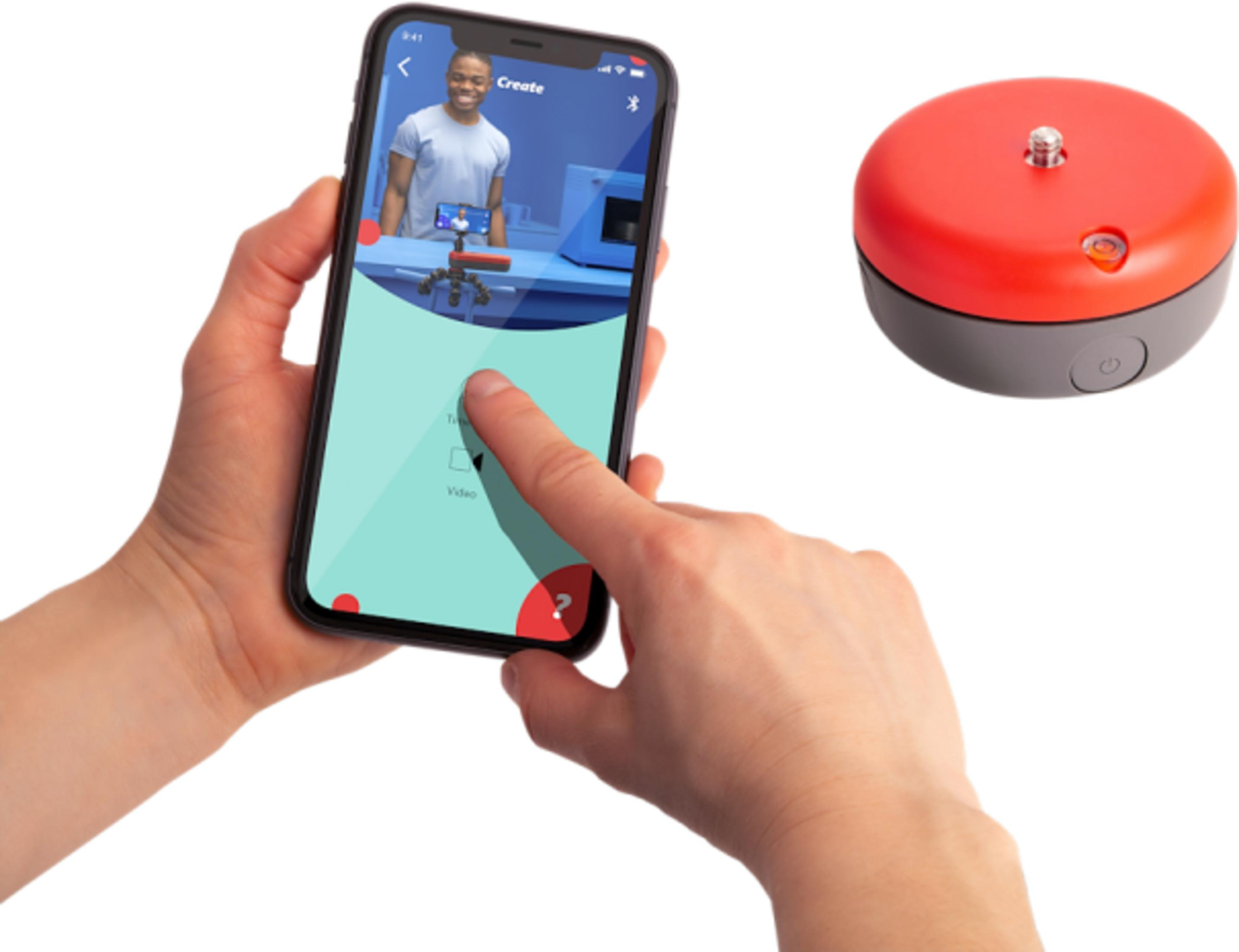 Eine Hand hält ein Smartphone, das eine App mit einem Mann zeigt. Daneben liegt ein rotes Gerät.