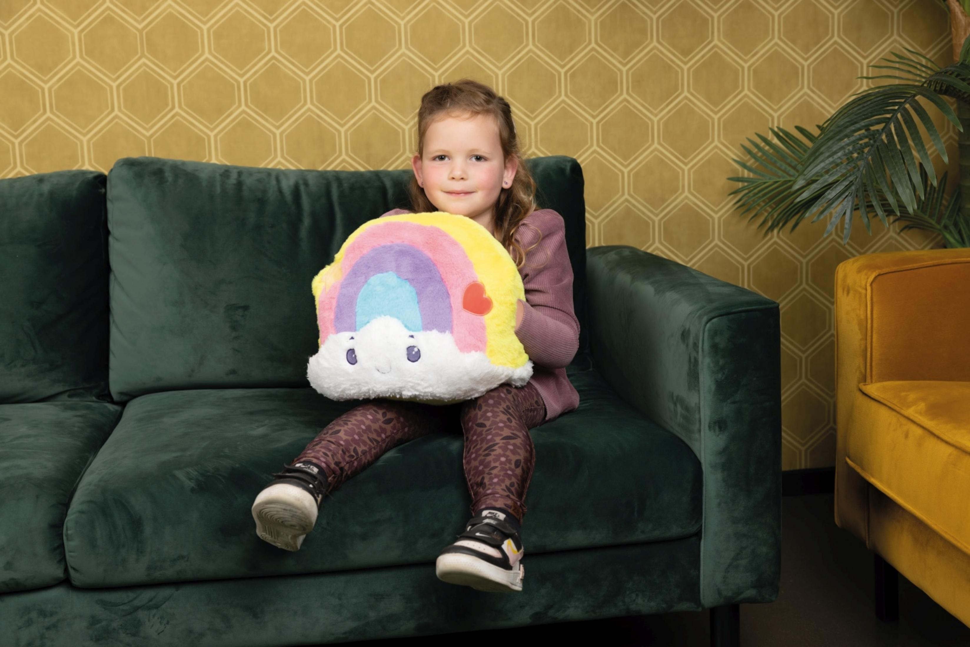 Ein junges Mädchen sitzt auf einer grünen Couch und hält ein buntes Regenbogn-Kissen.