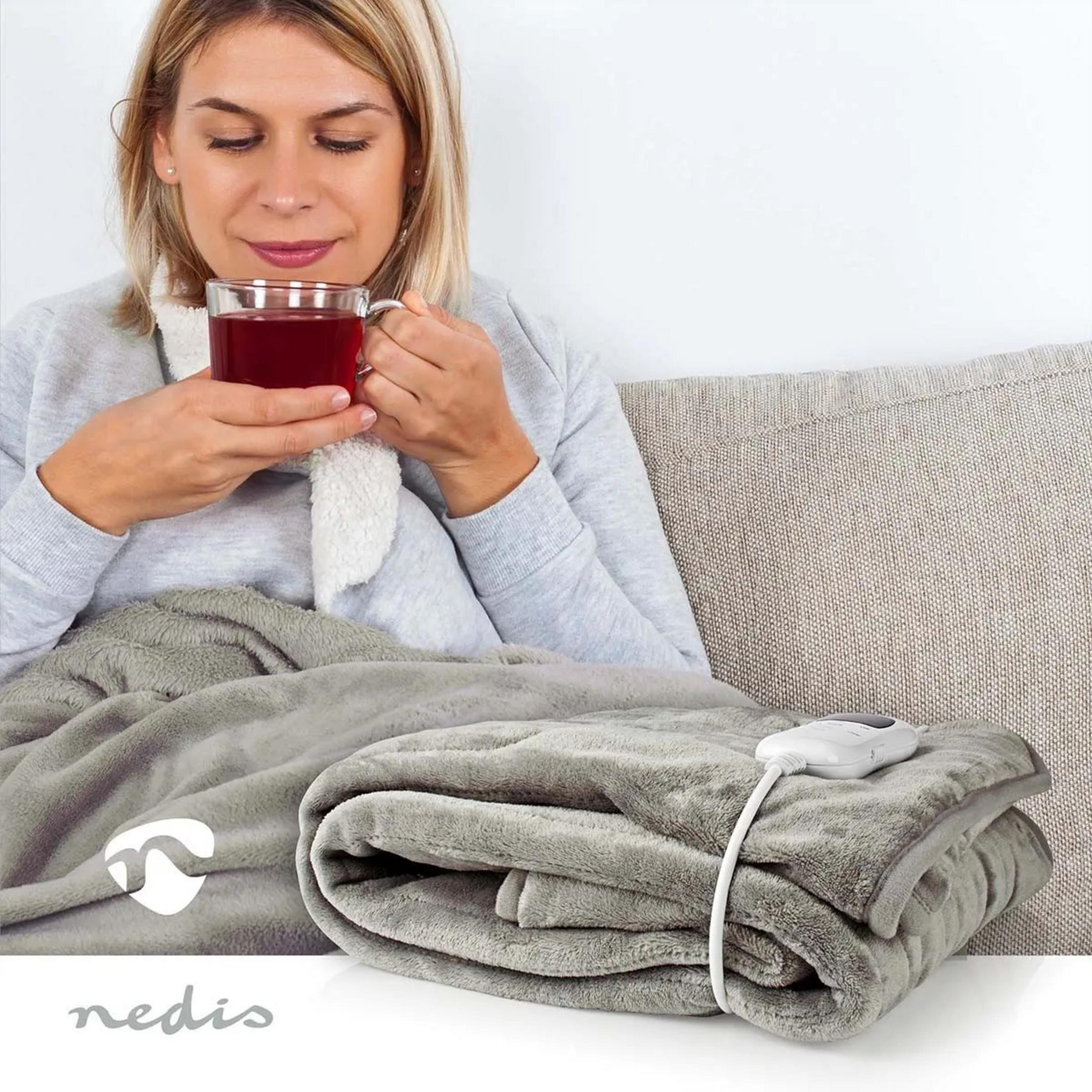 Kobieta pije herbatę, owinięta w szary koc elektryczny z pilotem.