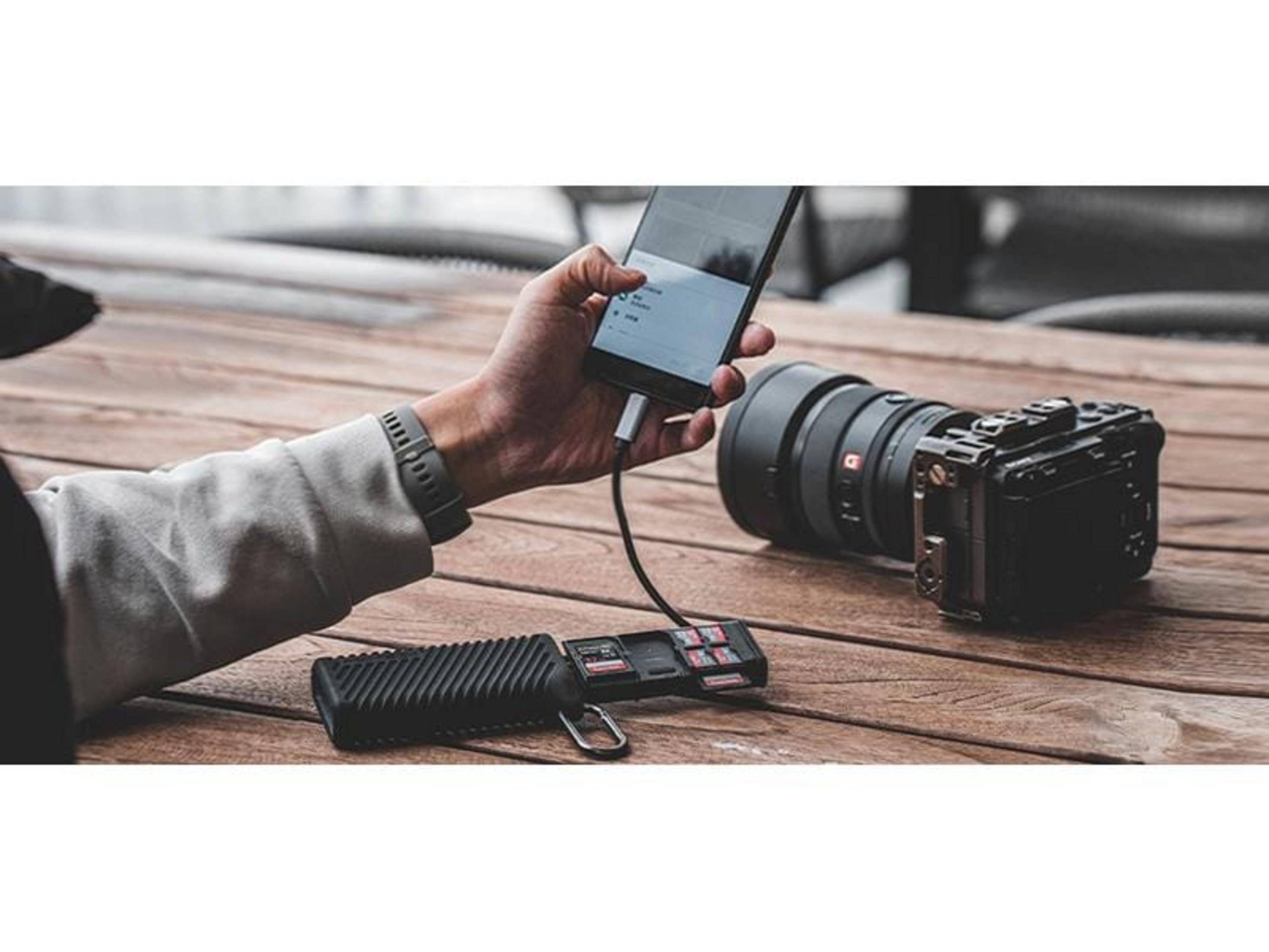 Dłoń trzyma smartfon podłączony do czarnego kabla. Obok leży aparat i czytnik kart.