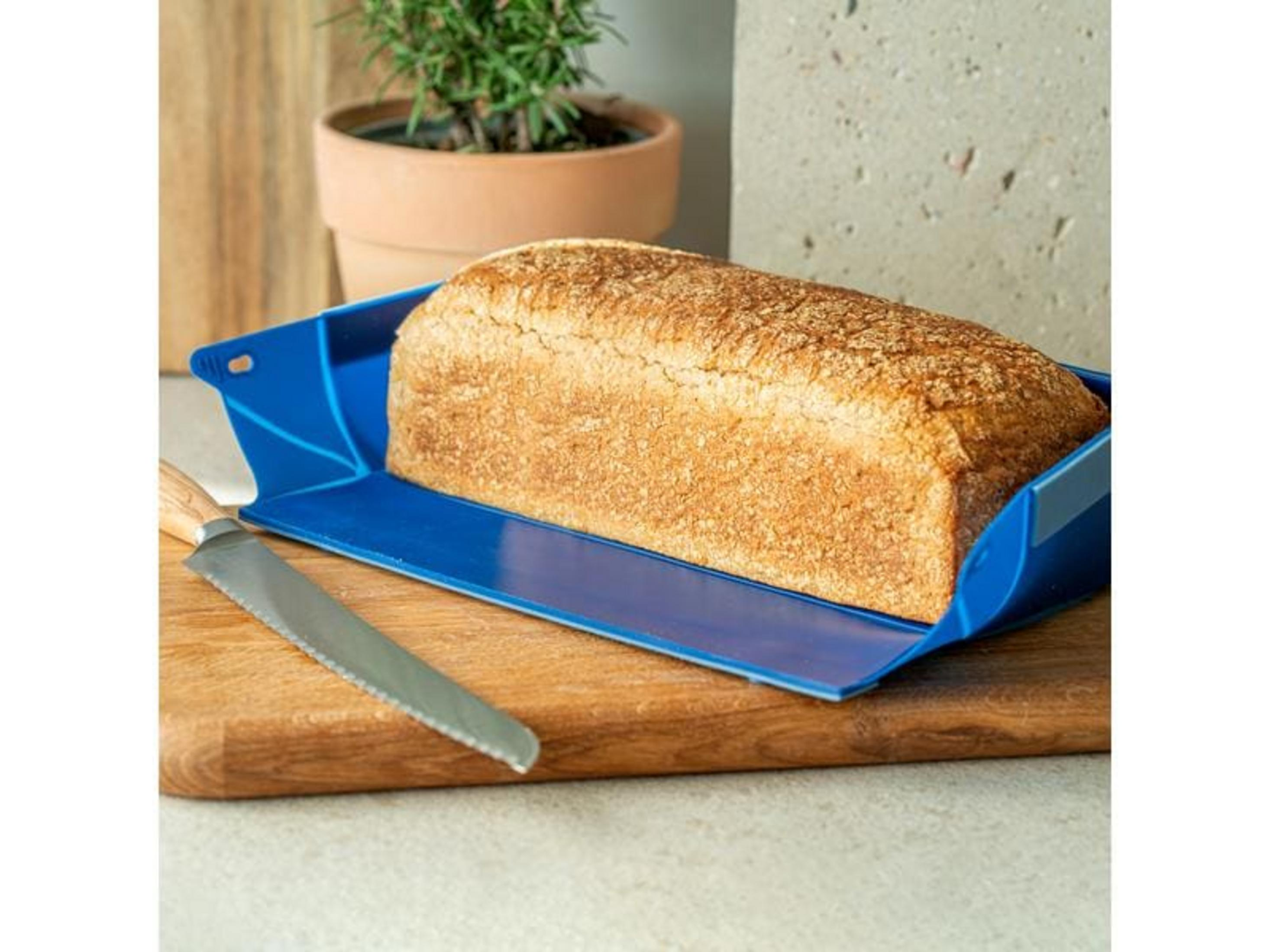 Ein goldbraunes Brot liegt in einem blauen Behälter auf einem Holzbrett neben einem Messer.