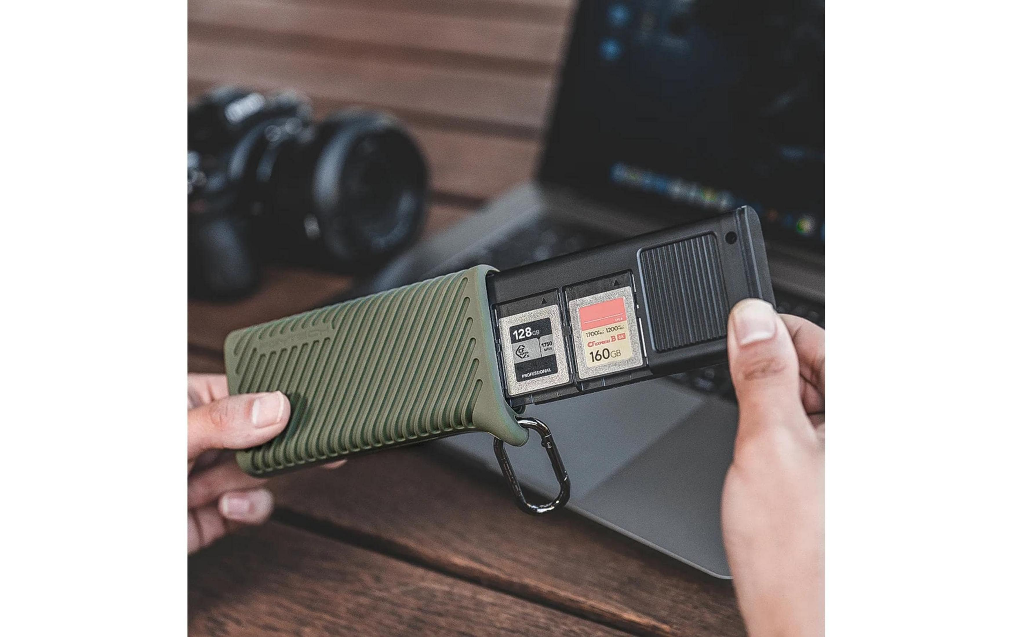 Dłonie trzymają zielone etui na karty pamięci z dwiema kartami, laptopem i aparatem w tle.