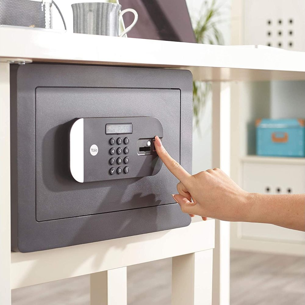 Una mano presiona el escáner de huellas dactilares de una caja fuerte gris montada bajo una mesa.