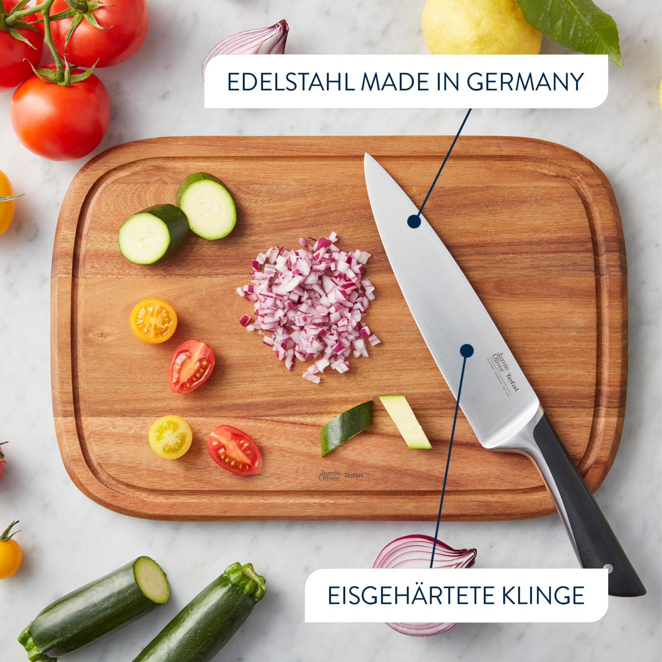 Ein Kochmesser liegt neben geschnittenen Zwiebeln, Tomaten und Zucchini auf einem Holzbrett.