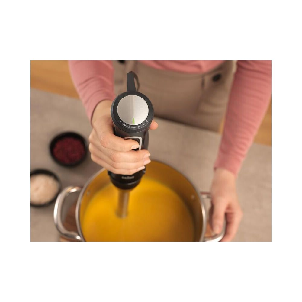 Ręka trzyma blender ręczny nad garnkiem z żółtą zupą.