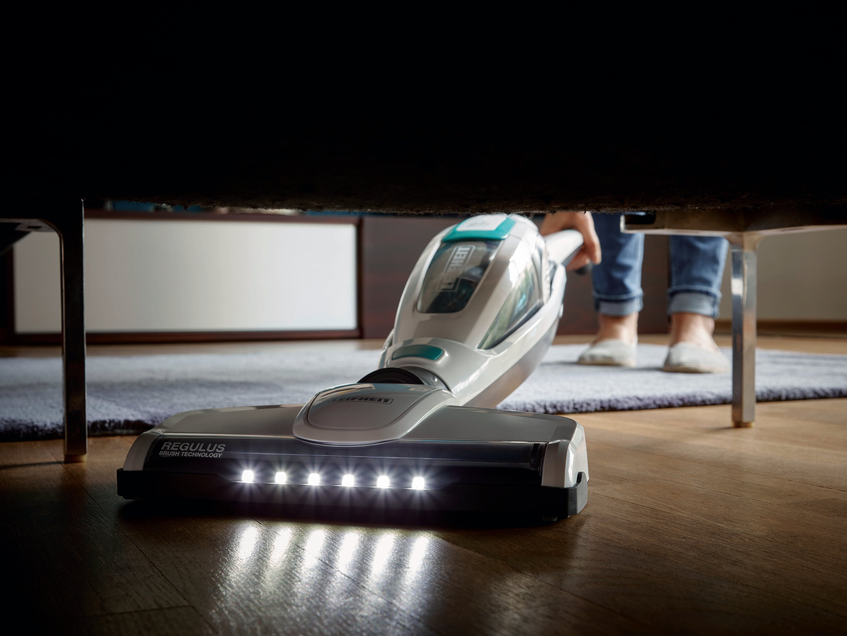 Odkurzacz bezprzewodowy ze światłami czyści drewnianą podłogę pod meblem.