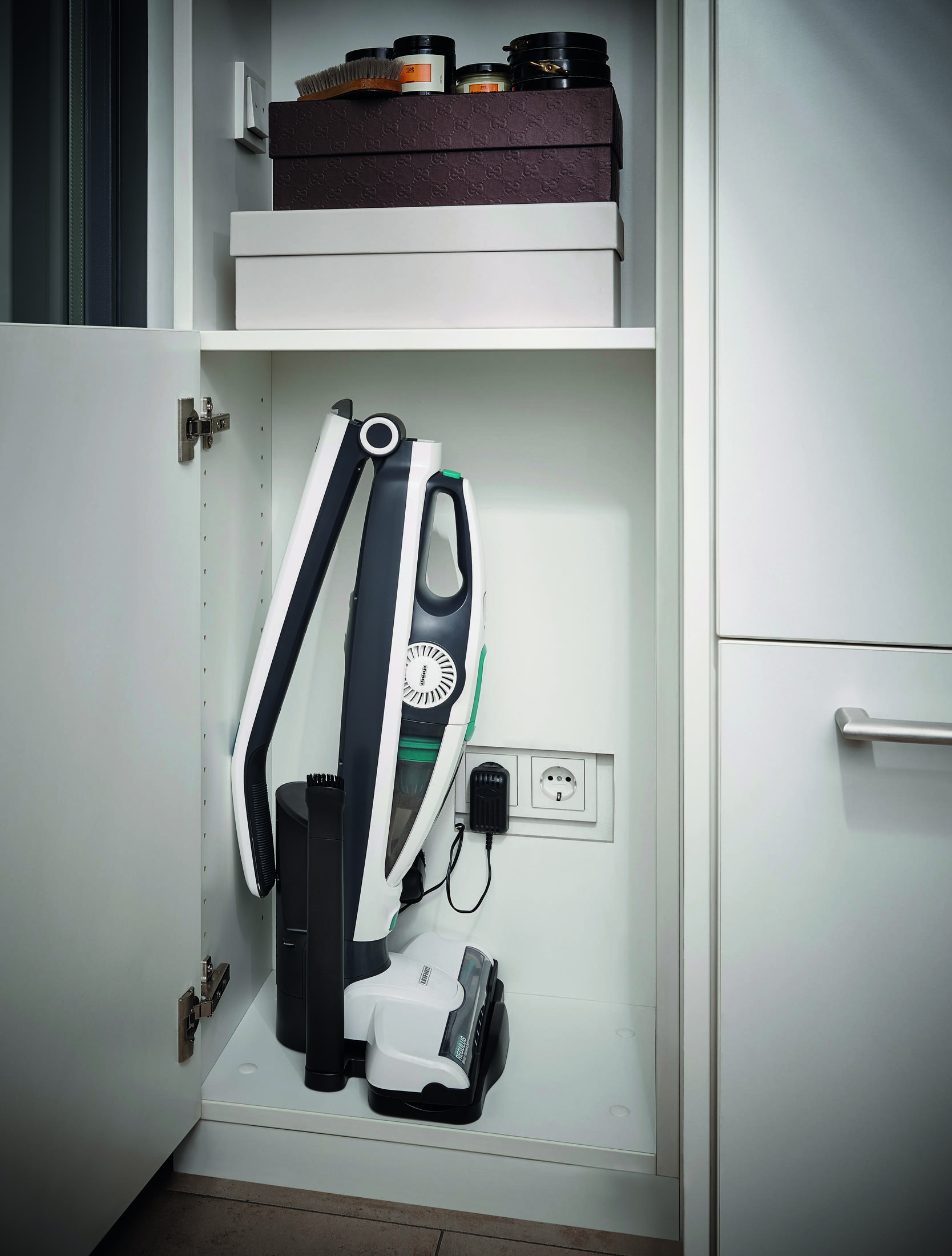 Biała szafka z otwartą półką. W środku stoi odkurzacz.