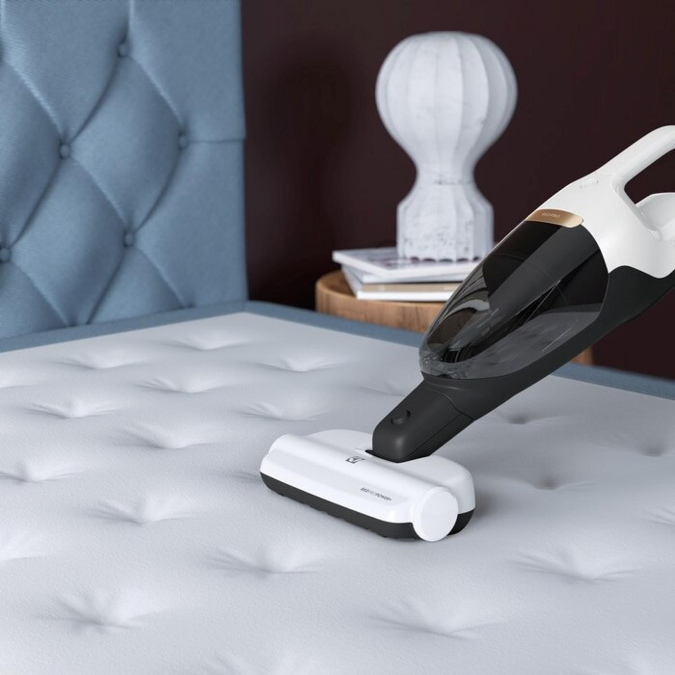 Un aspirateur blanc portatif nettoie un matelas blanc à côté d'une tête de lit bleue.